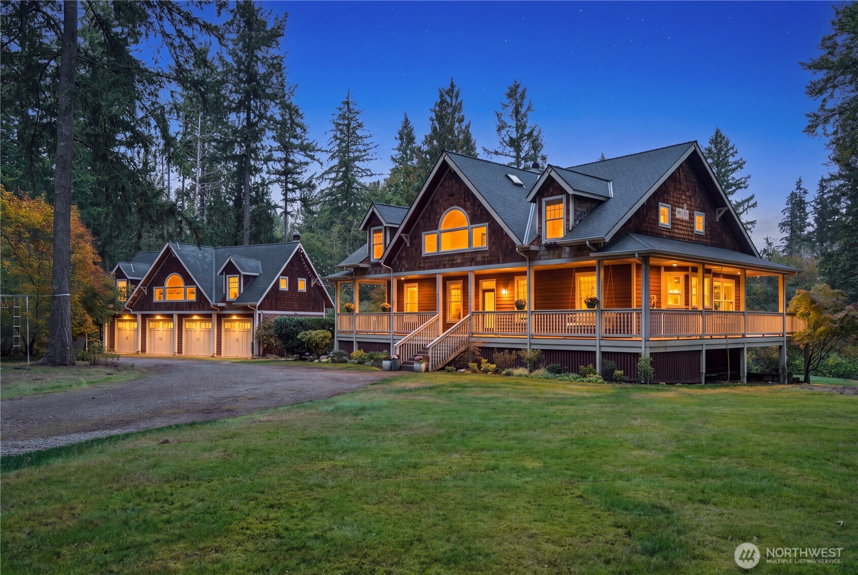 Wood-shingle home with a wraparound porch and detached three-car garage, warmly lit at dusk.