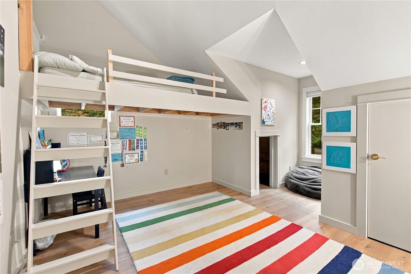 Bedroom with a white loft bed, desk underneath, vaulted ceiling, and colorful striped rug.