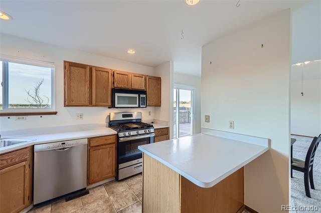 Kitchen with wood cabinets, stainless-steel appliances, and a white-topped peninsula beside windows and a deck door.