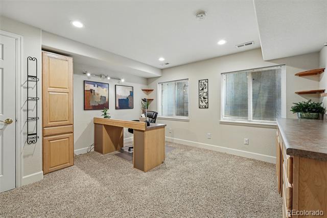 Home office with a wooden desk, beige carpet, and countertop beneath windows with blinds.