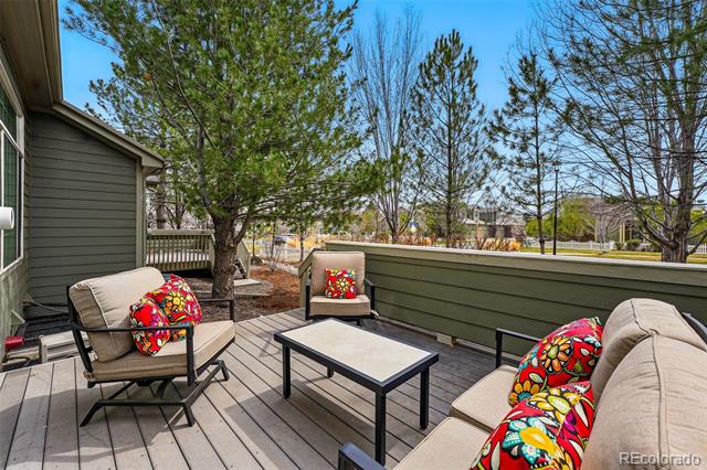 Deck with cushioned seating and colorful pillows, enclosed by a low railing beneath mature trees.