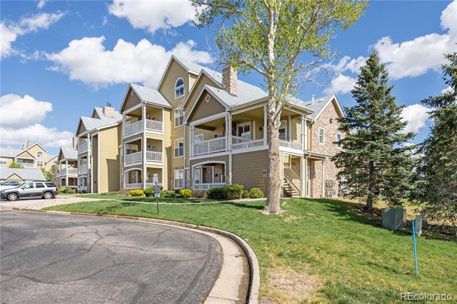 Condominium building with multiple white-railed balconies beside a curved driveway and mature trees.