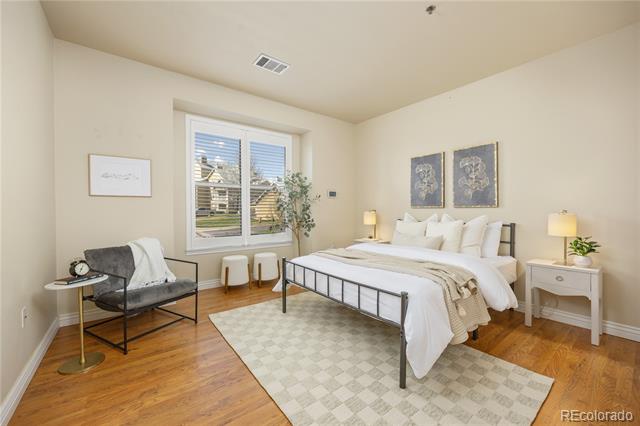 Staged bedroom with white bedding, a black metal bed, hardwood floors, and a large window overlooking homes.