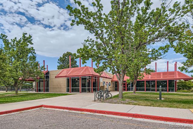Commercial building with a red metal roof, glass frontage, and bicycle racks beneath mature trees.