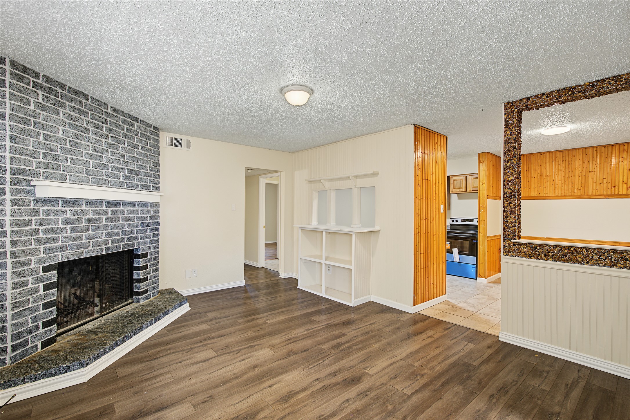 Empty living room with a gray brick fireplace, wood-look floors, and an opening to the kitchen.