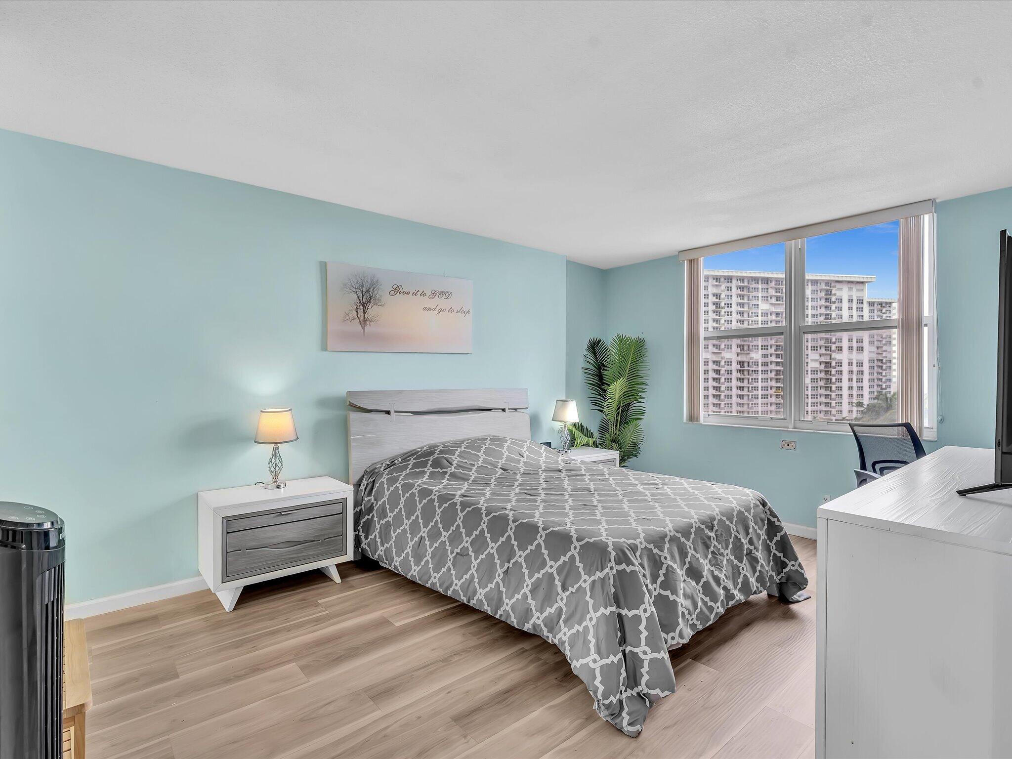 Bedroom with a gray patterned bedspread and a large window overlooking a neighboring high-rise.