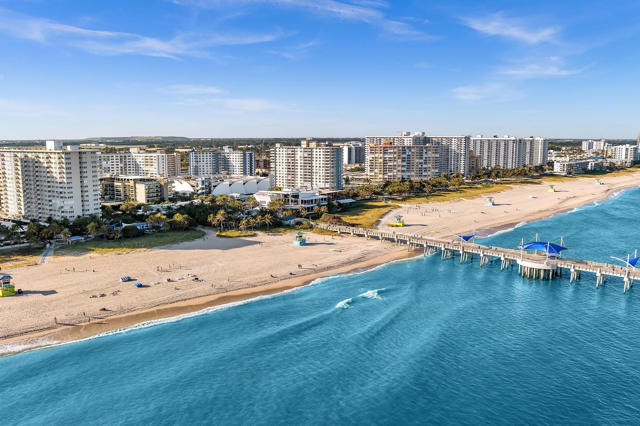 Aerial view of Pompano Beach, with beachfront towers, a sandy shore, and a pier extending over turquoise water.