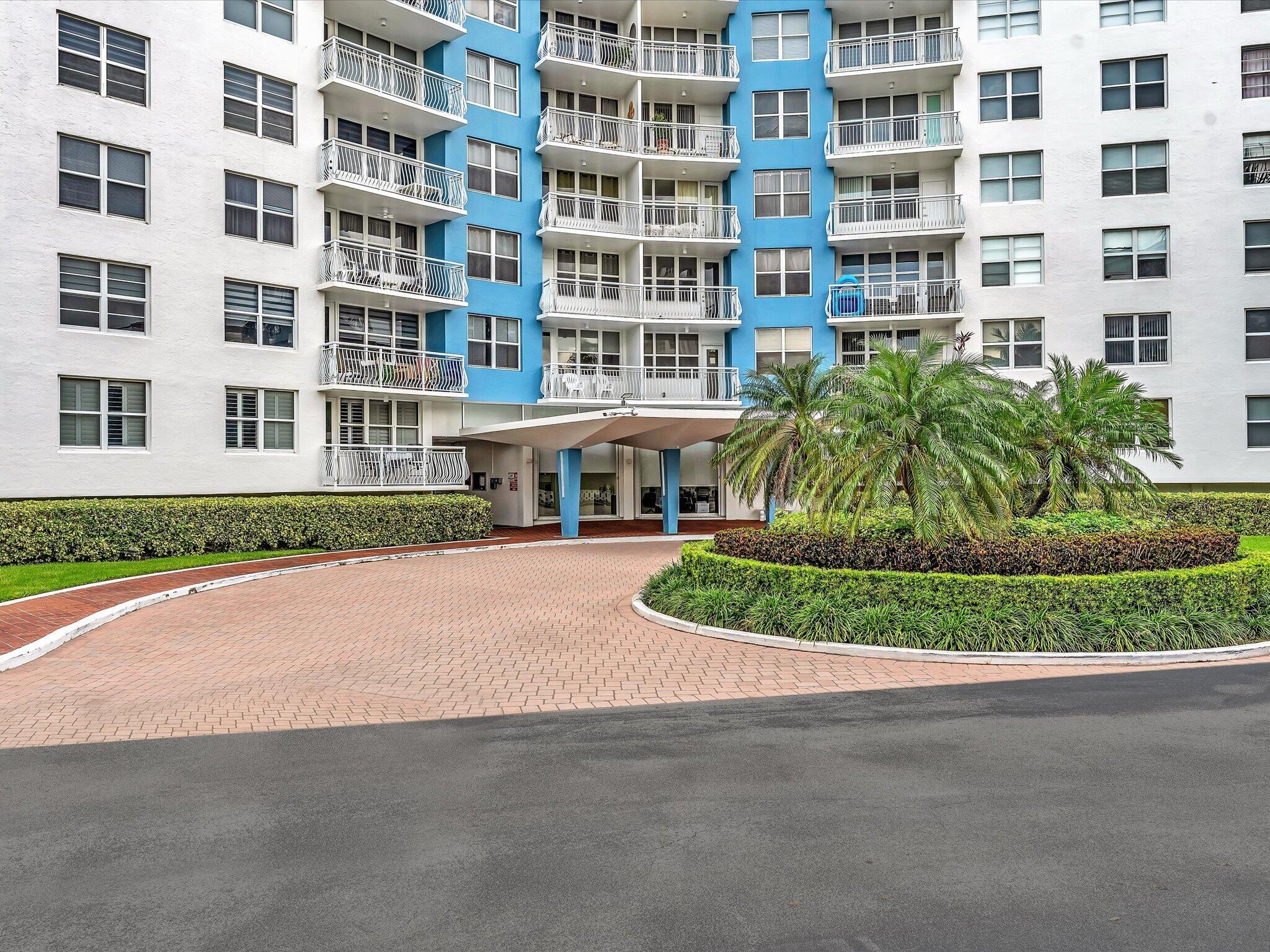 White and blue condominium building with balconies, palm trees, and a brick-paved entrance driveway.
