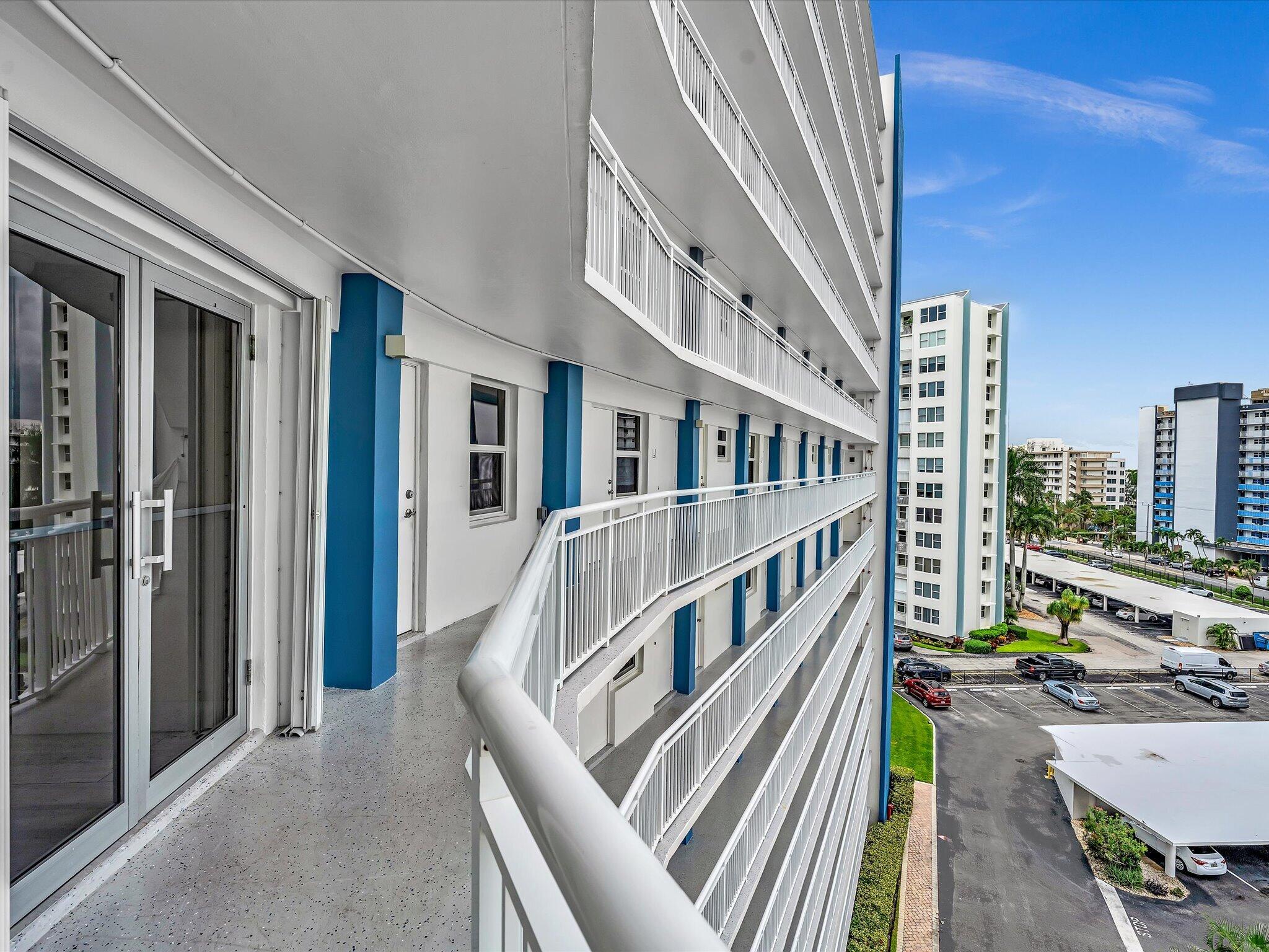 Condominium balcony corridor with white railings and blue accents overlooking nearby towers and parking.