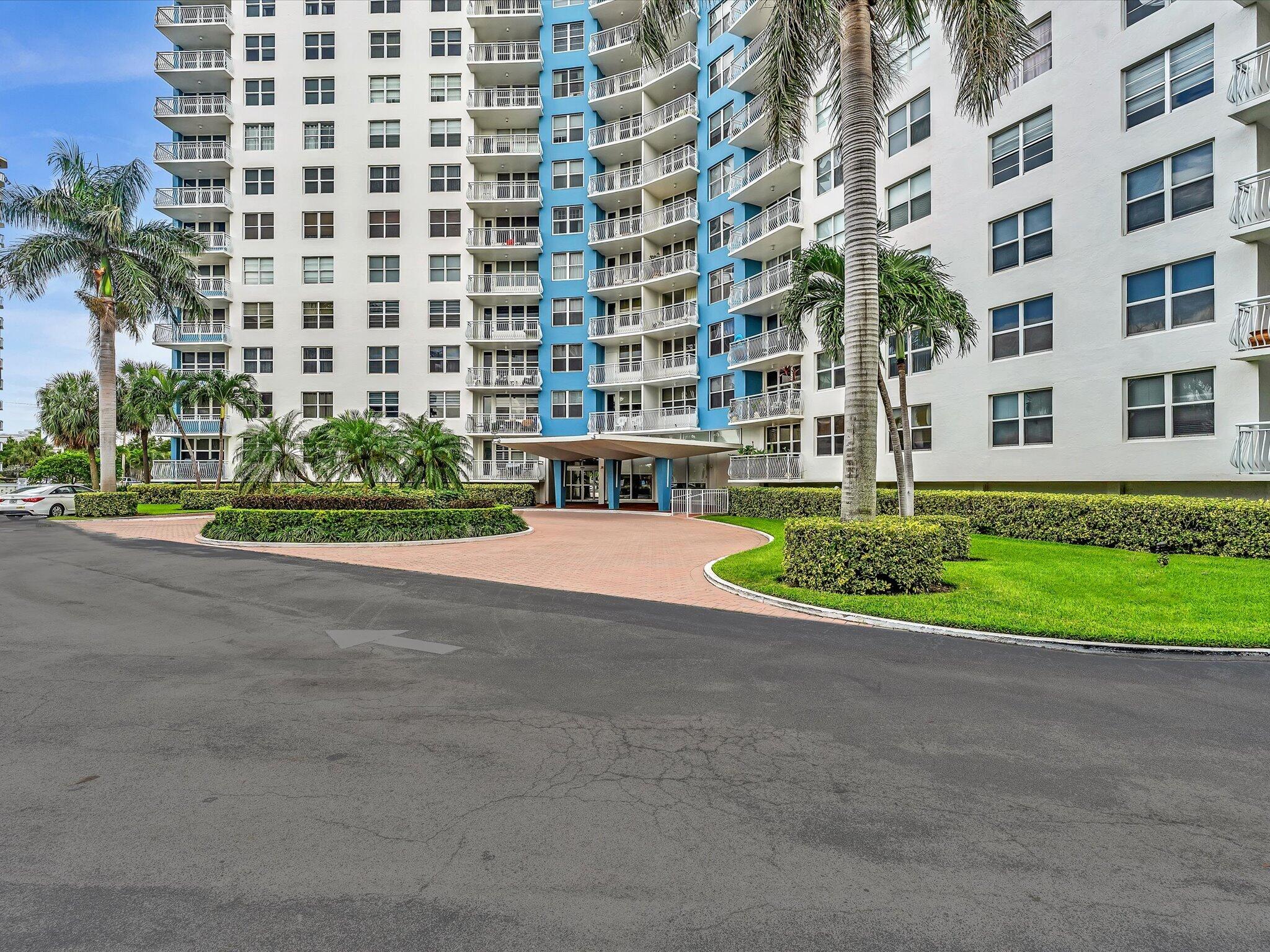 White and blue condominium entrance with balconies, palm trees, and a curved brick driveway.