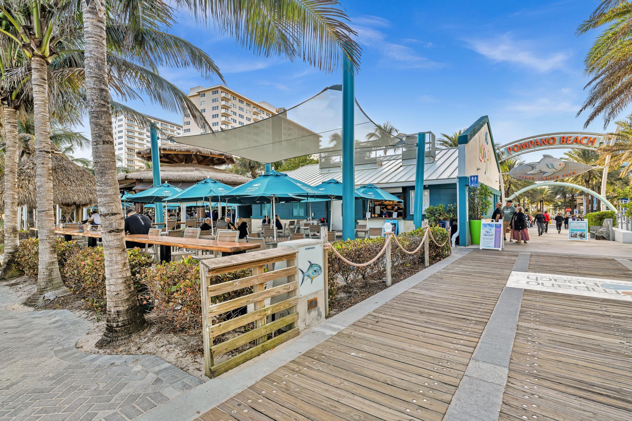 Pompano Beach Fisher Family Pier entrance beside a palm-lined boardwalk and outdoor restaurant.