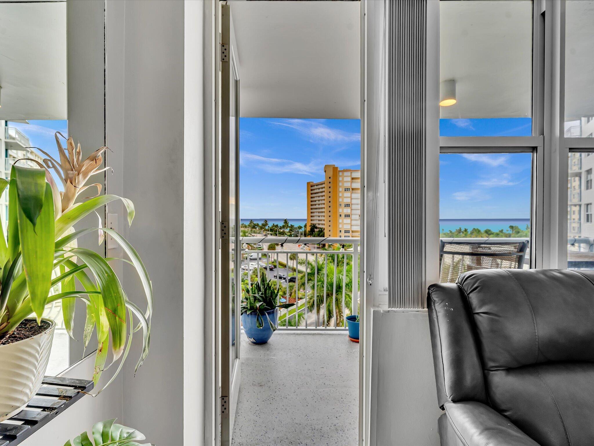 Living room opens onto a balcony overlooking the ocean, with potted plants and a nearby high-rise.