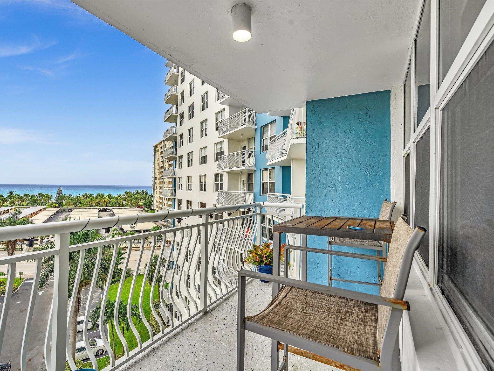 Condominium balcony with a table and chairs overlooking palm trees and the ocean.