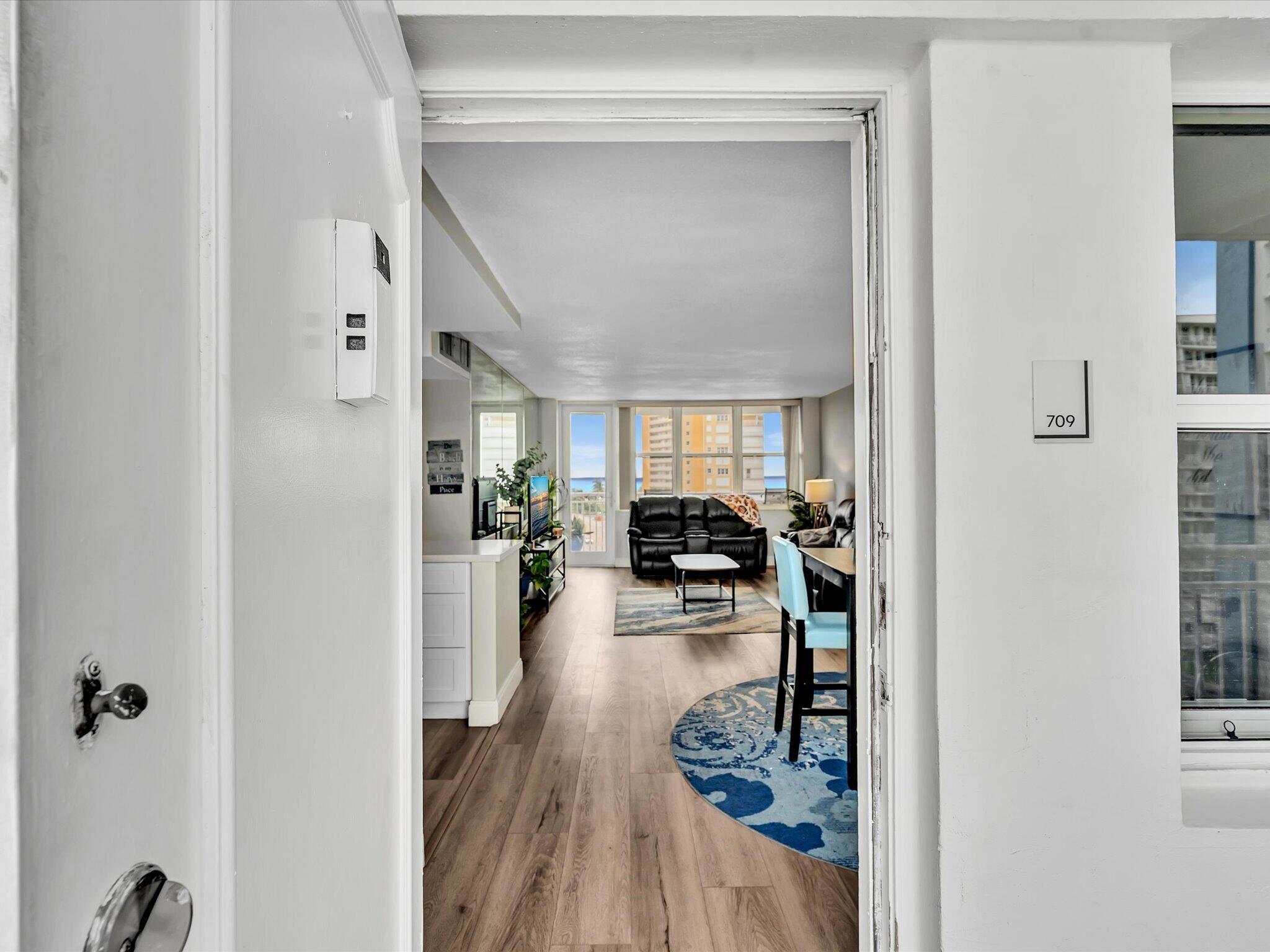 Condo entrance opening onto a furnished living room with ocean glimpses through large windows.