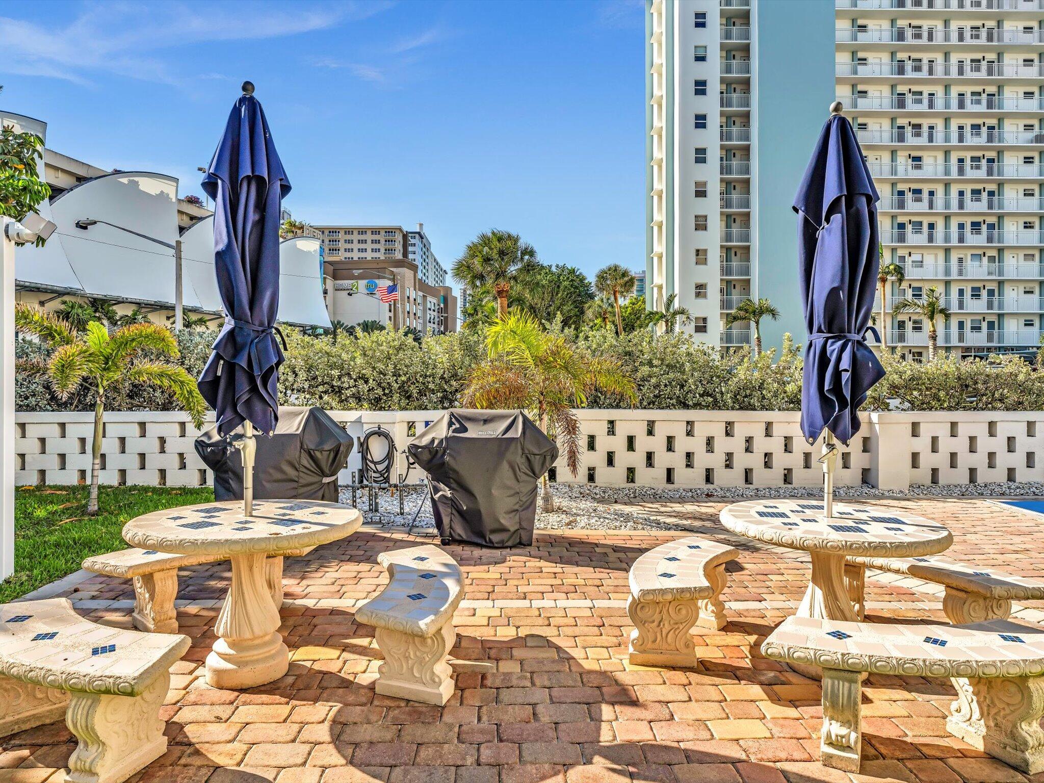 Outdoor courtyard with stone picnic tables, closed umbrellas, covered grills, and tropical landscaping.