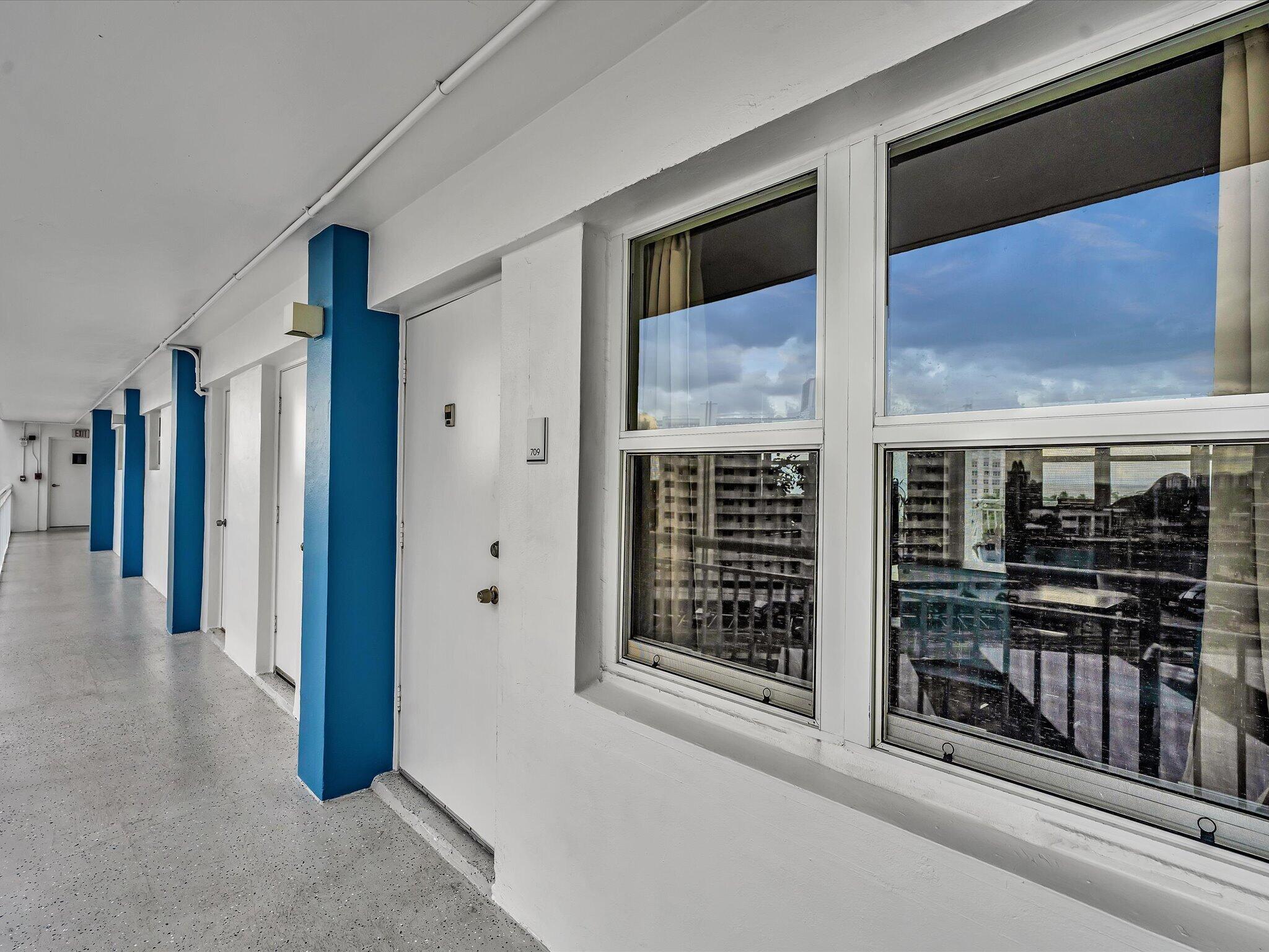 Condominium hallway with blue columns, unit doors, and windows overlooking neighboring buildings.