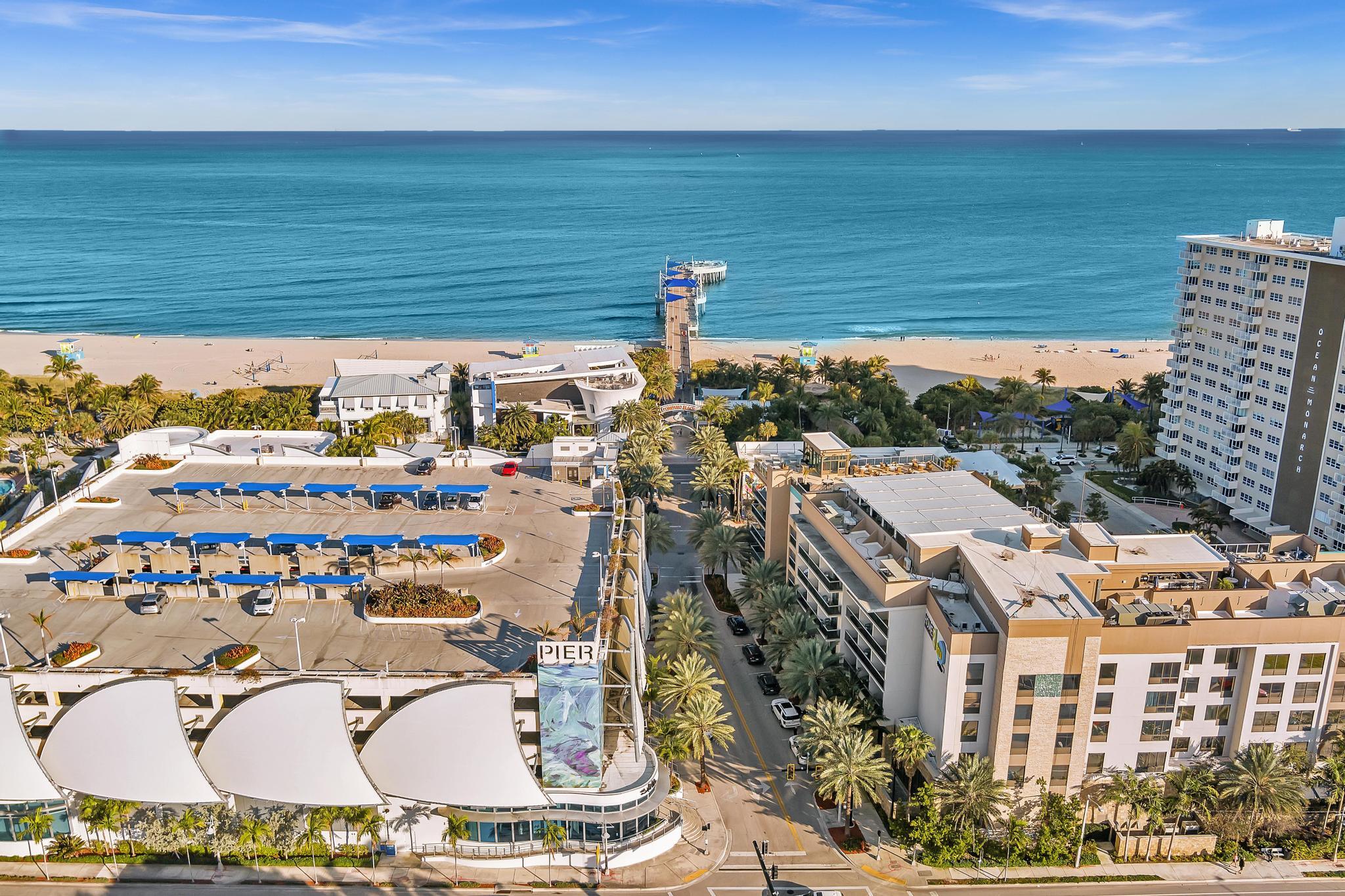 Aerial view of beachfront buildings, a pier, sandy shore, and turquoise ocean.