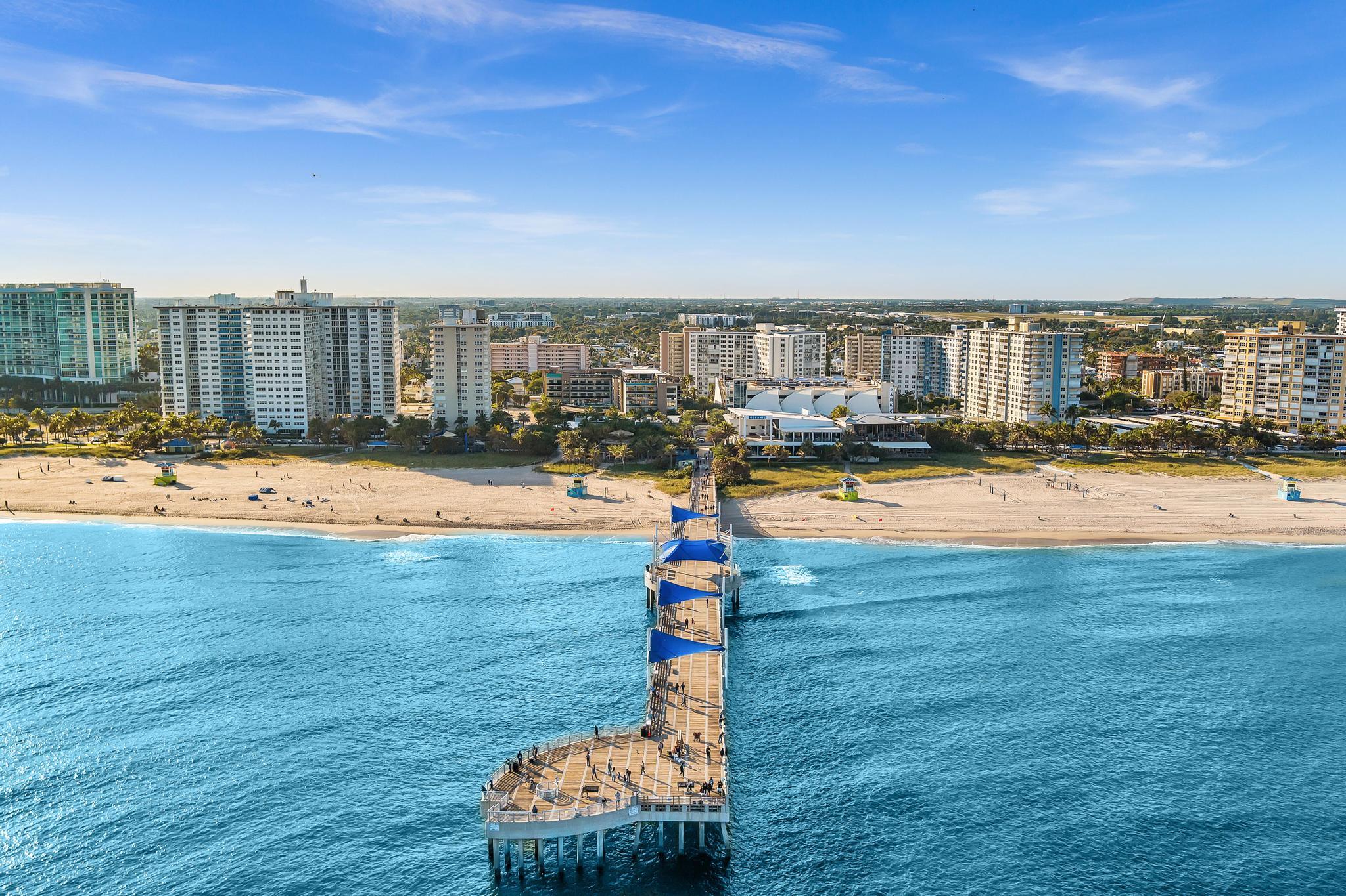 Aerial view of a beachfront pier extending into turquoise water, with sandy shore and oceanfront towers.