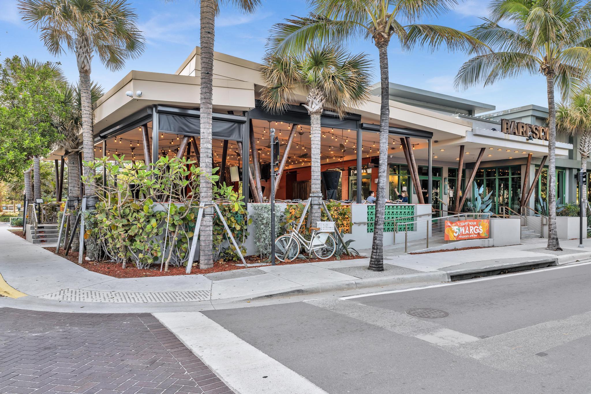 Modern glass-front restaurant with a string-lit patio, tropical landscaping, and palm trees.