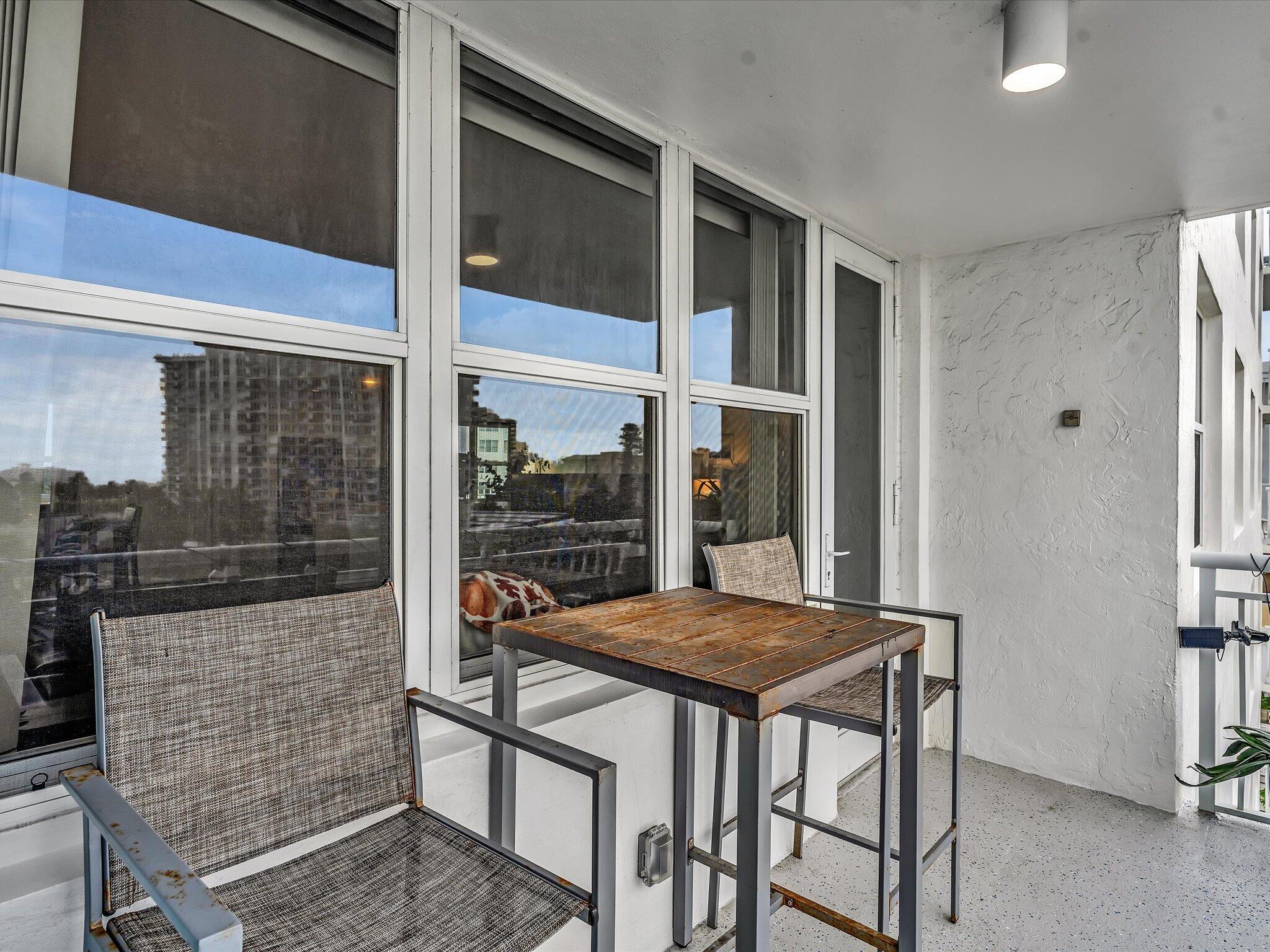 Apartment balcony with a wooden table, woven chairs, and large windows reflecting nearby buildings.