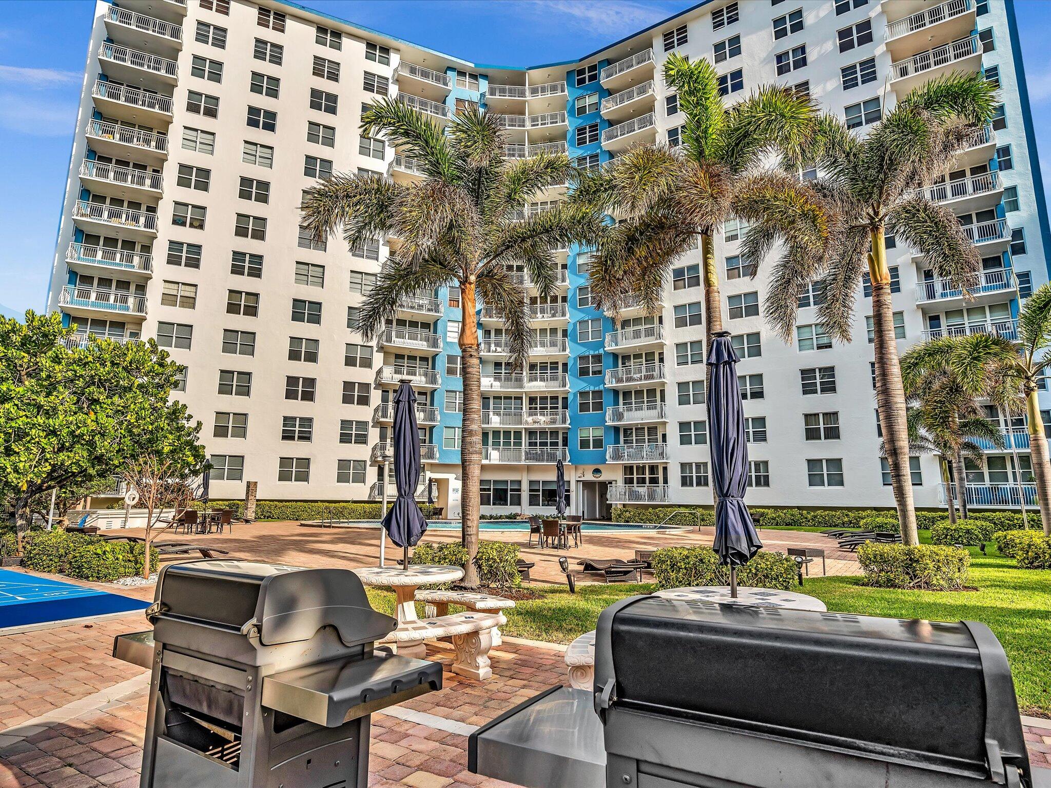 Condominium courtyard with palm trees, a pool, and barbecue grills in front of a white-and-blue building.