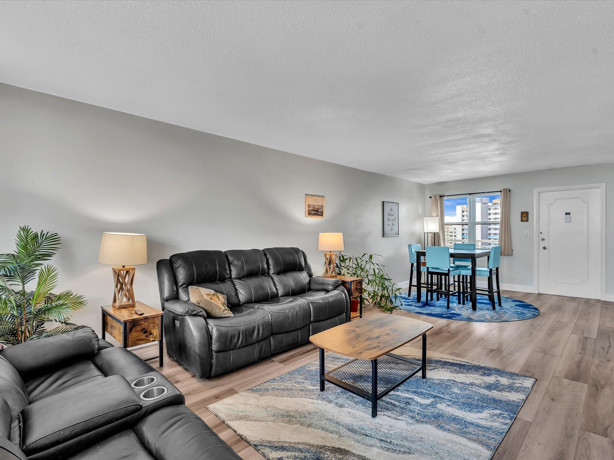 Living room with black leather sofas and a dining area beside a window overlooking high-rise buildings.