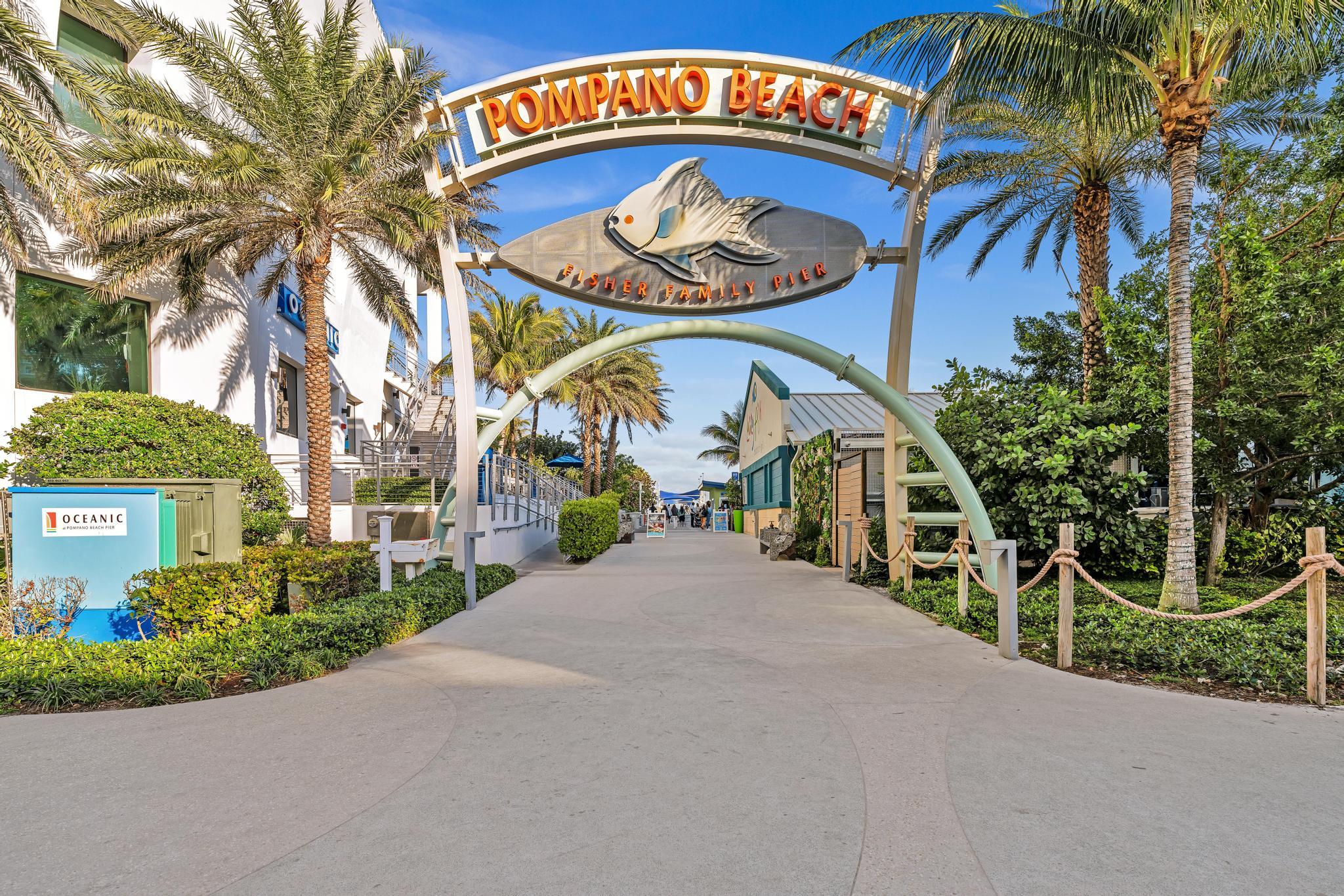 Fisher Family Pier entrance beneath the Pompano Beach arch, lined with palm trees and greenery.
