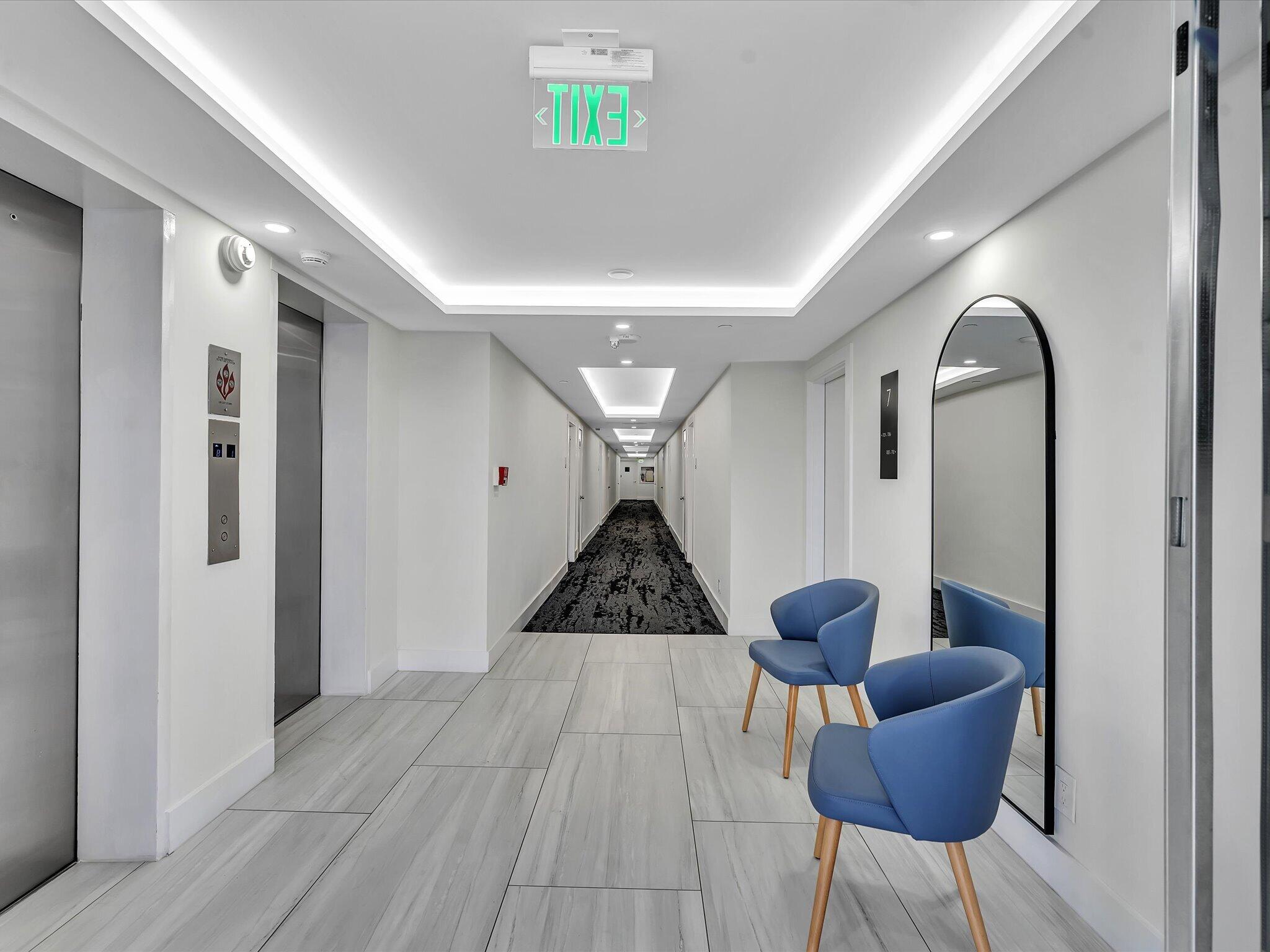 Modern condominium hallway with elevator doors, blue chairs, and a tall arched mirror.