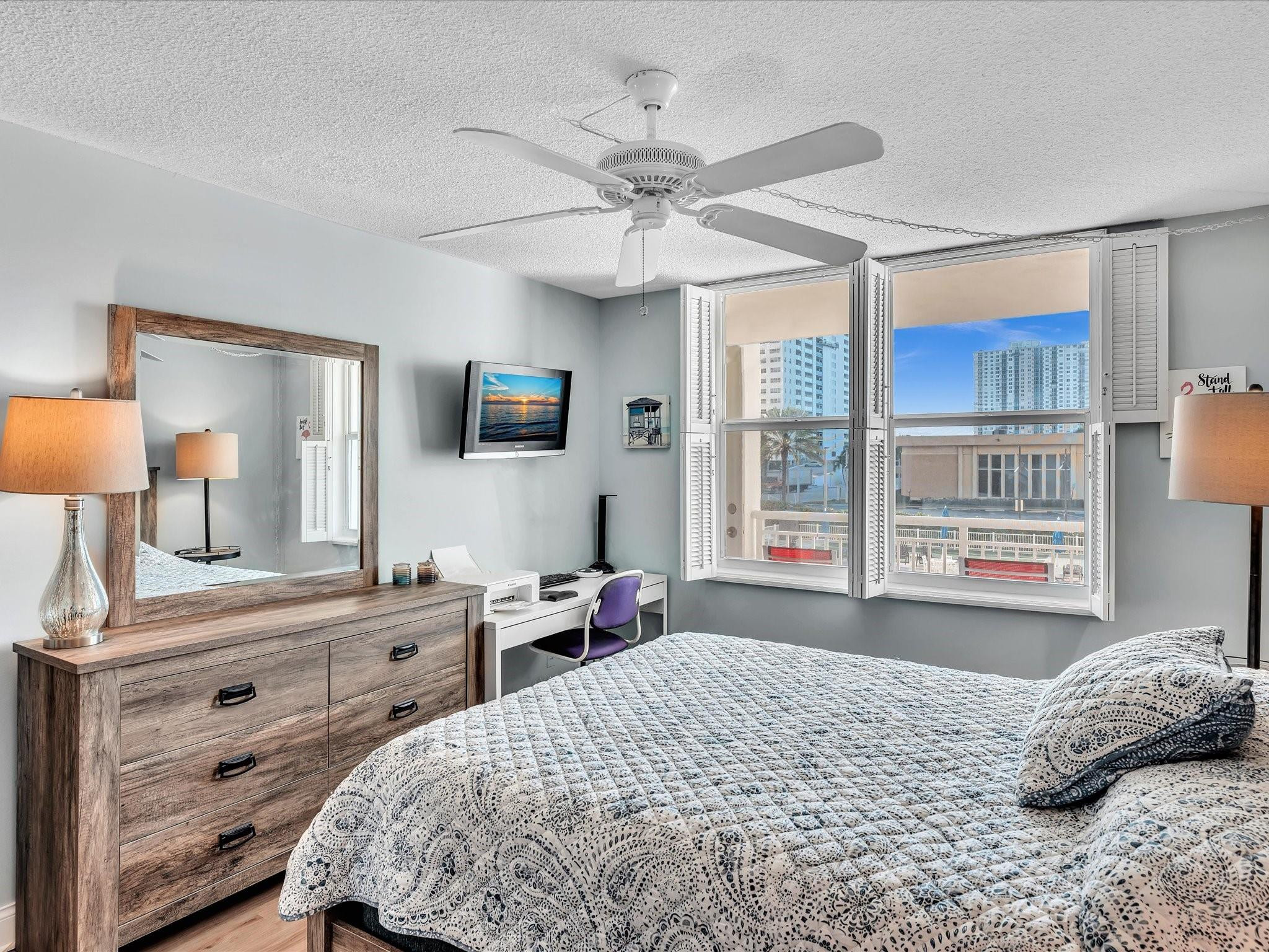 Bedroom with shuttered windows, a patterned bedspread, wood dresser, and desk.
