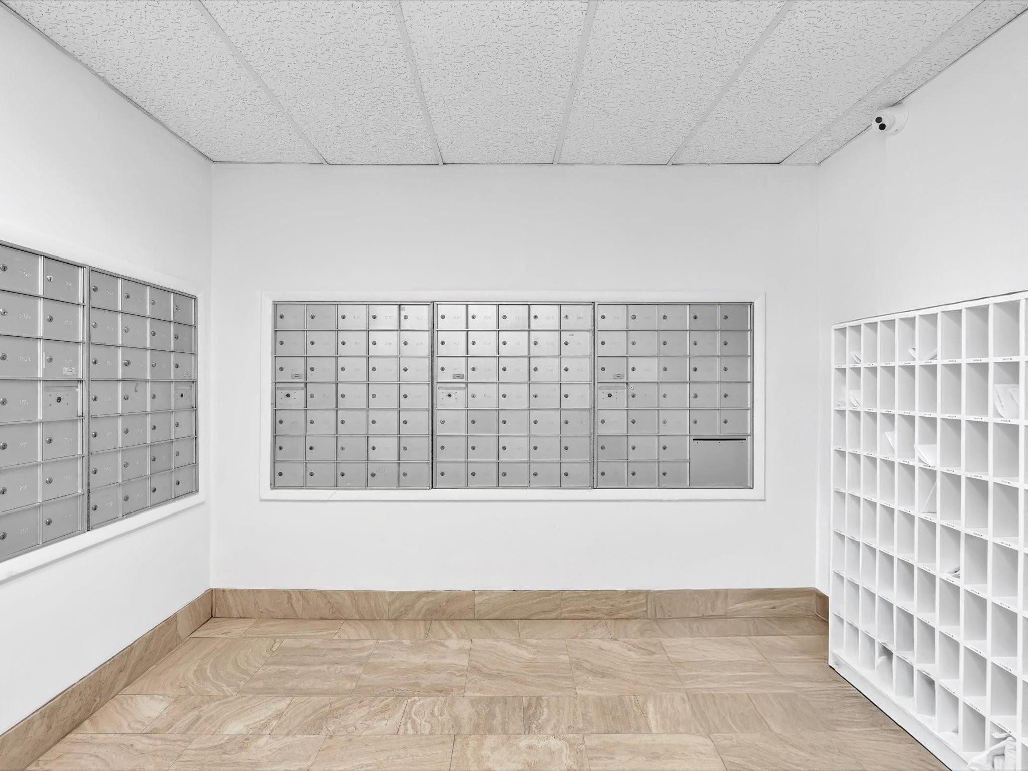 Condominium mailroom with metal mailboxes, white parcel cubbies, and beige stone tile flooring.