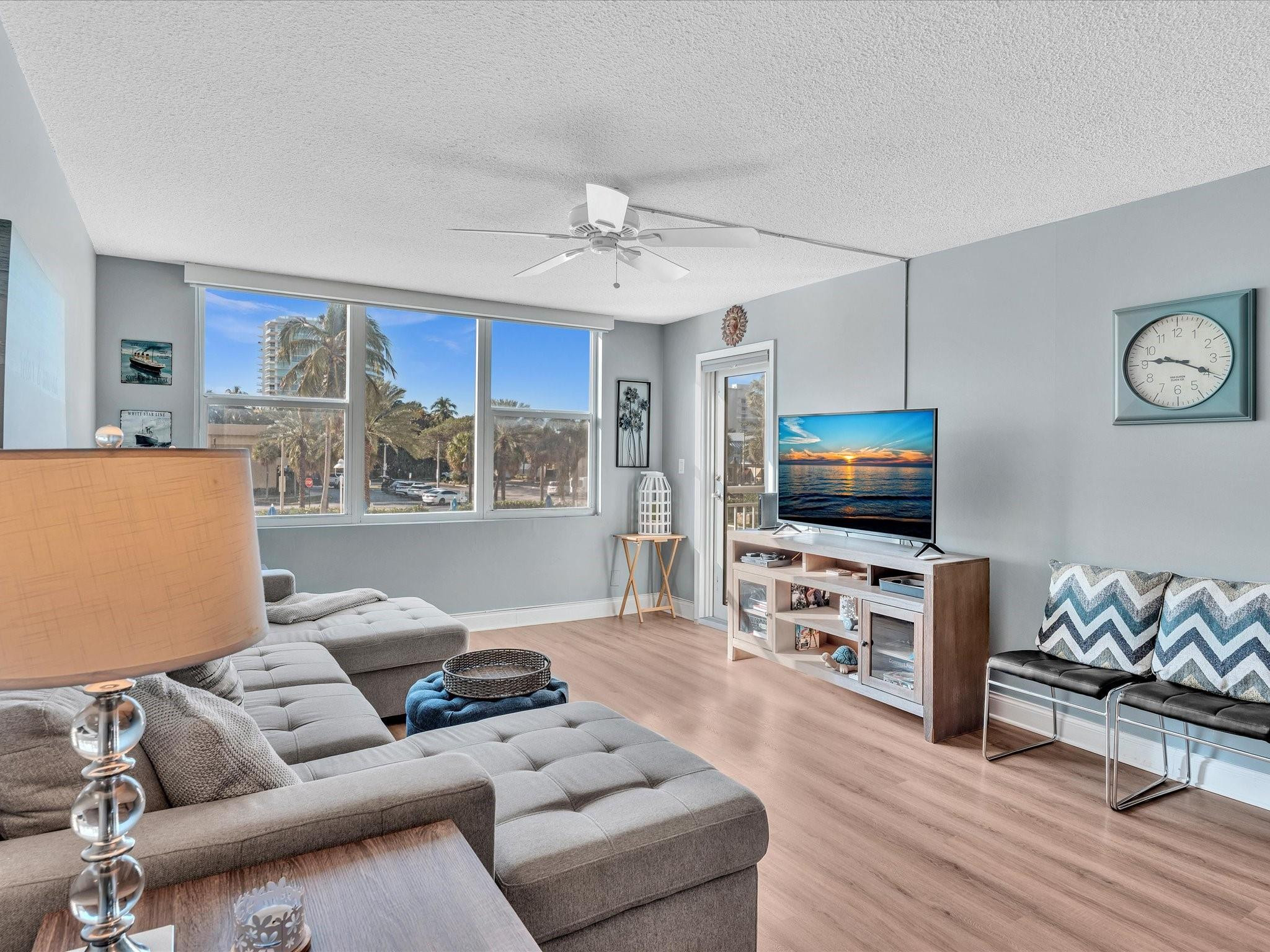 Living room with a gray sectional sofa and wide windows overlooking palm trees.