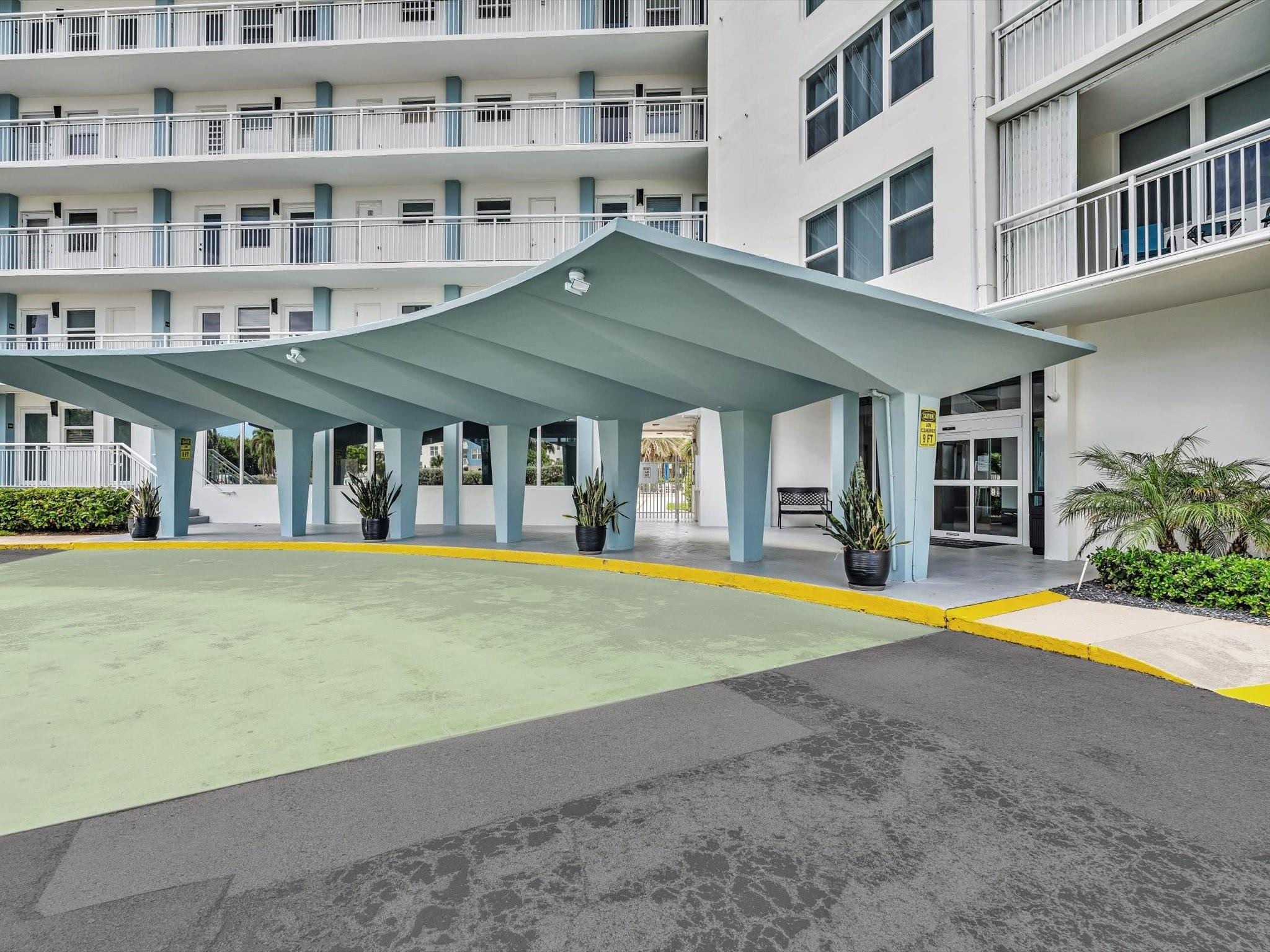 Condominium entrance beneath a broad pale blue porte-cochère, with balconies and potted plants.
