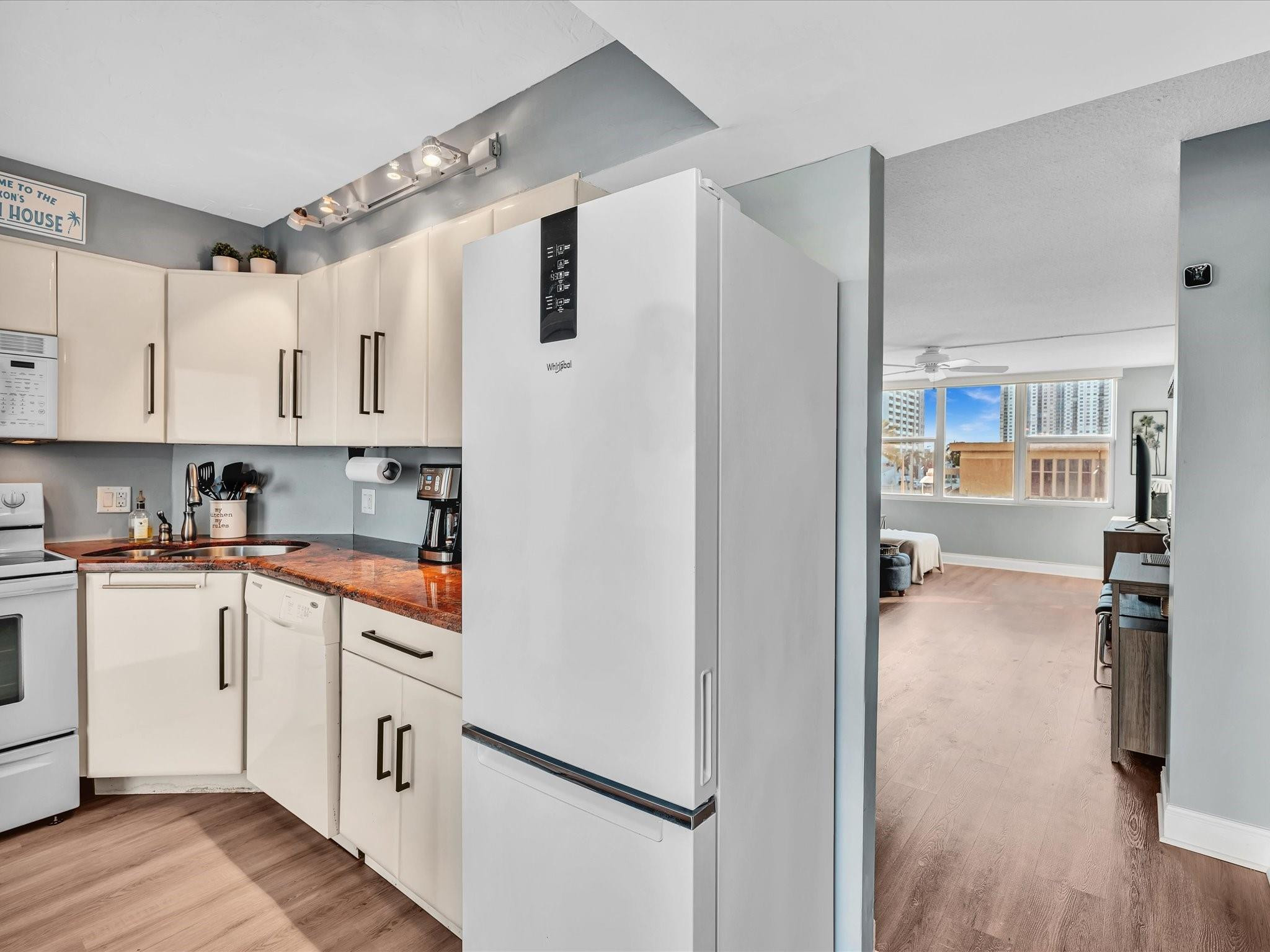 Condominium kitchen with white cabinetry, reddish stone counters, and a large refrigerator beside the living area.