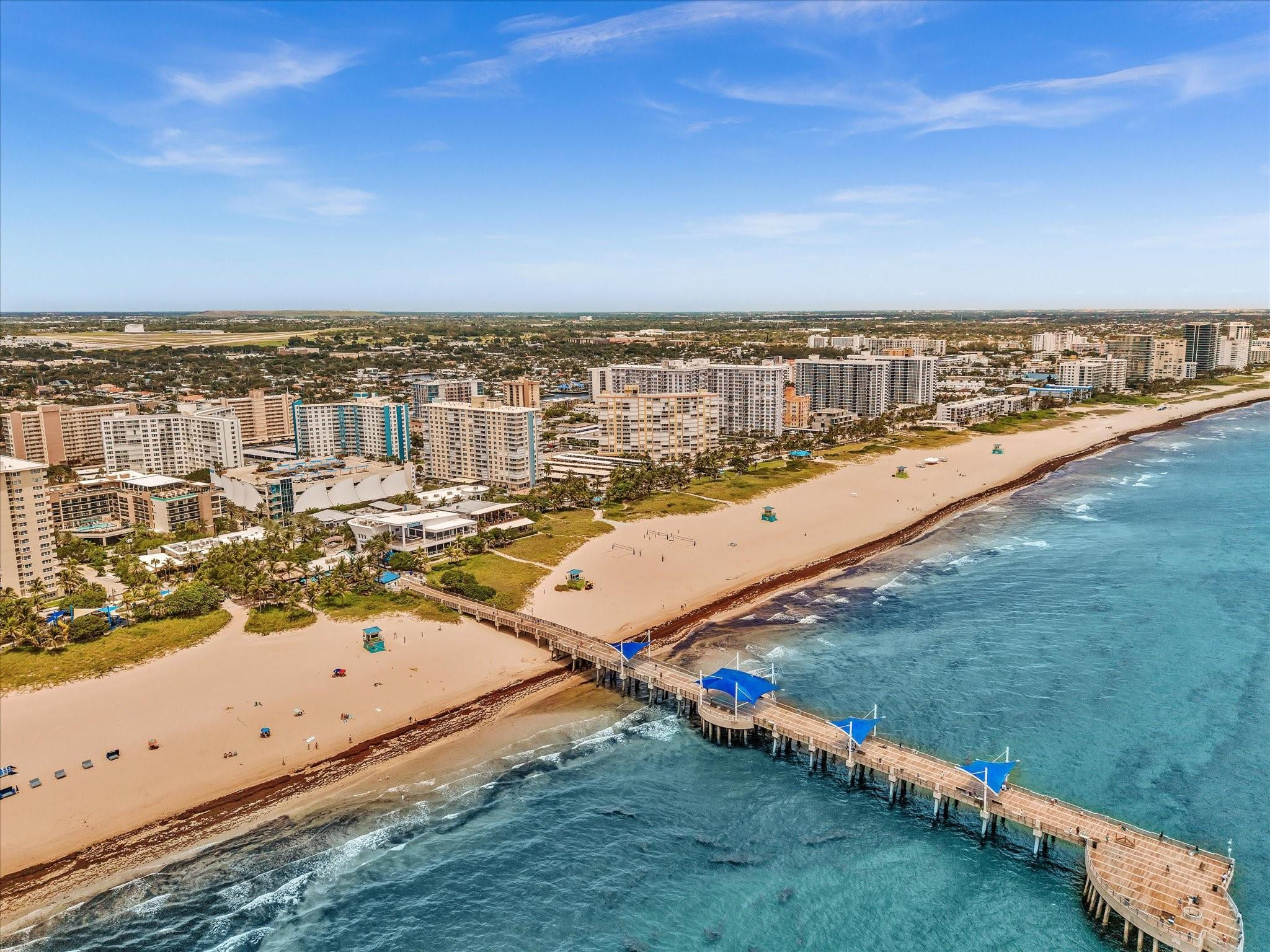 Aerial coastline with beachfront towers, sandy beach, and a long pier with blue canopies.