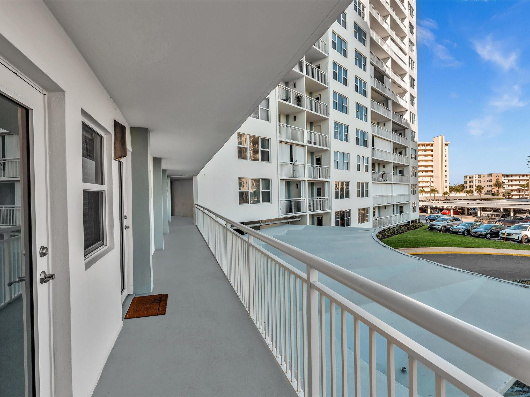 Condominium balcony walkway with white railings overlooking neighboring high-rises and a parking area.