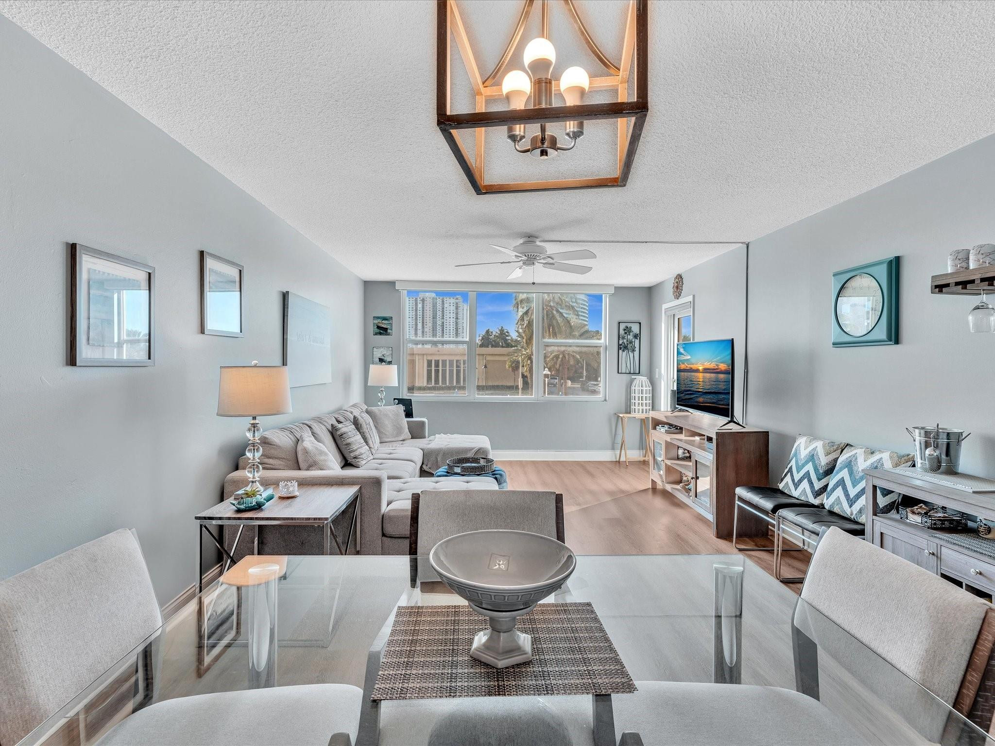 Furnished condo living room with a gray sectional and dining table beside large windows overlooking palm trees.