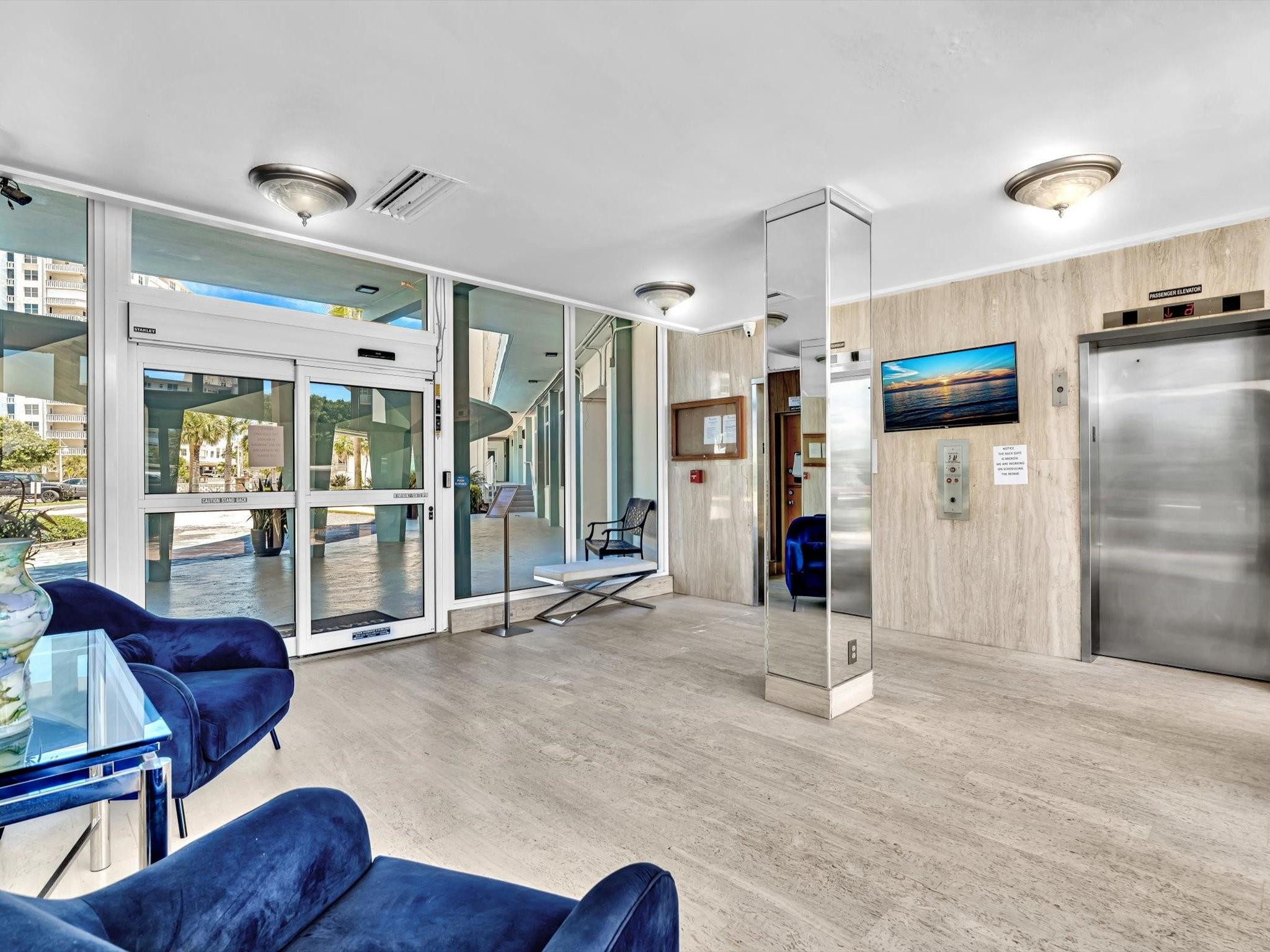 Condominium lobby with a glass entrance, mirrored column, elevator, and blue seating.