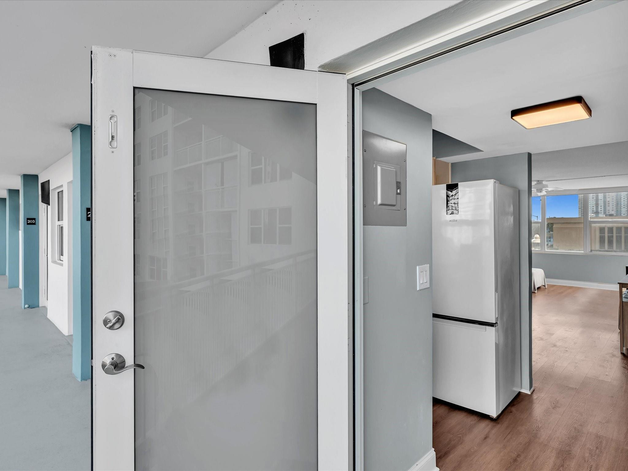Open screened condo door beside a refrigerator, with a wood-floored room visible beyond.