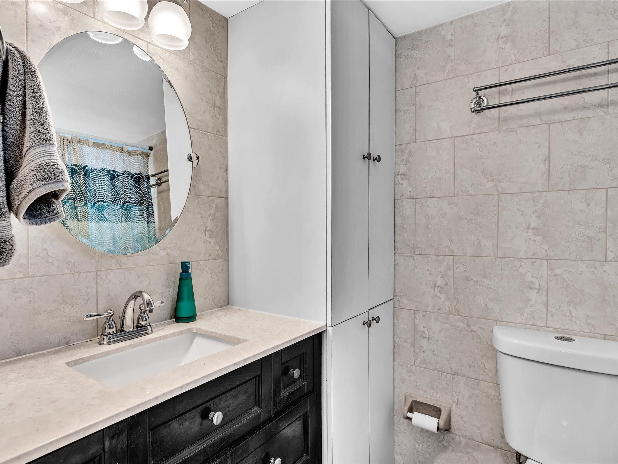Bathroom with a dark wood vanity, oval mirror, and light stone tile walls.