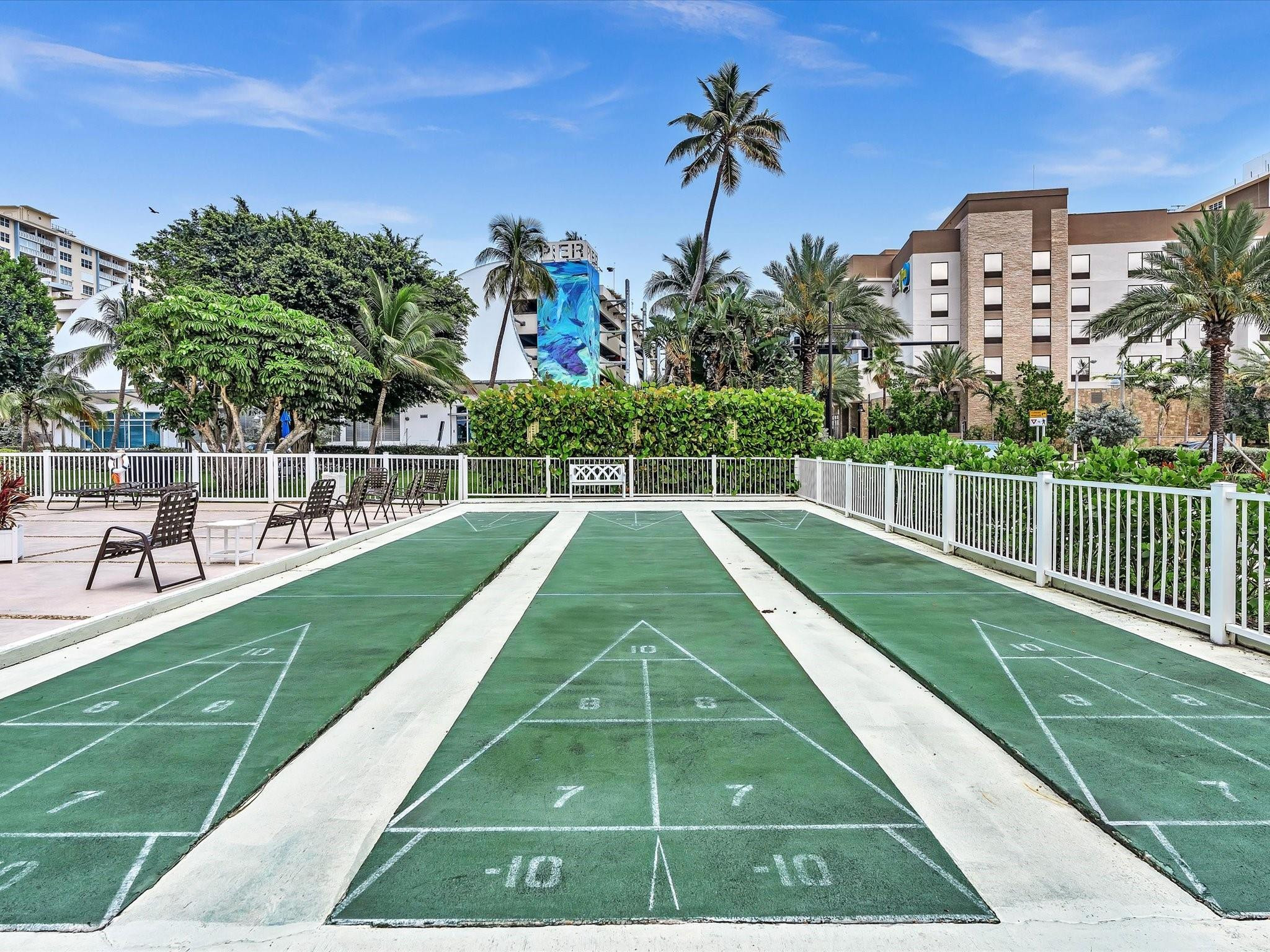 Outdoor shuffleboard courts with green numbered lanes, surrounded by palm trees and condo buildings.