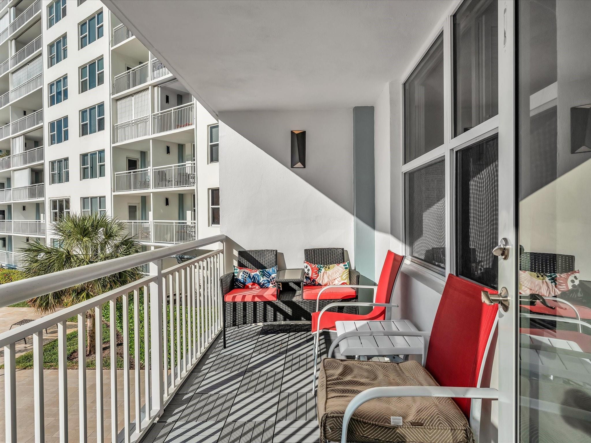 Condo balcony furnished with red-cushioned chairs, overlooking a neighboring building and palm trees.