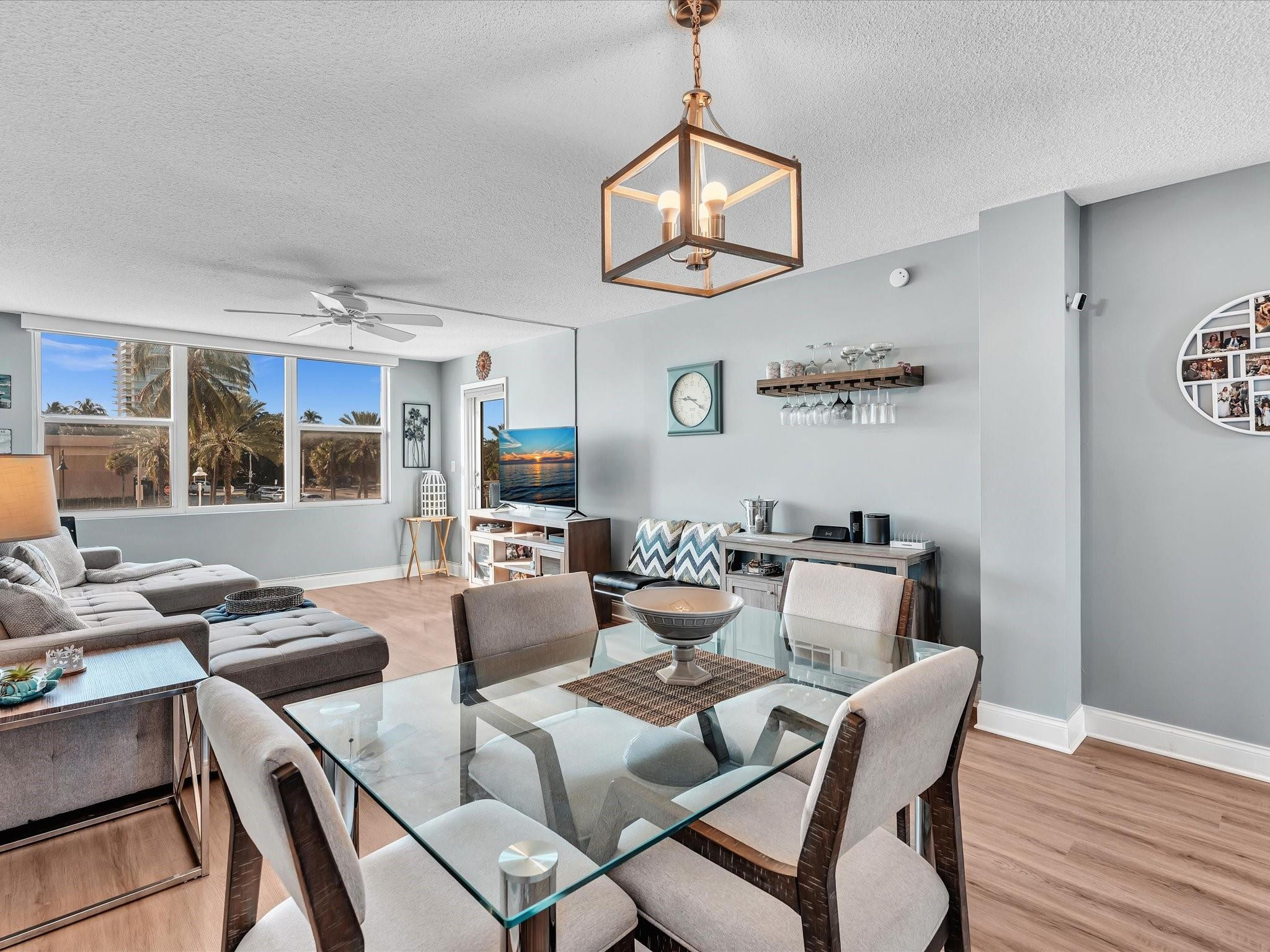 Furnished living and dining room with a glass table, sectional sofa, and large windows overlooking palm trees.