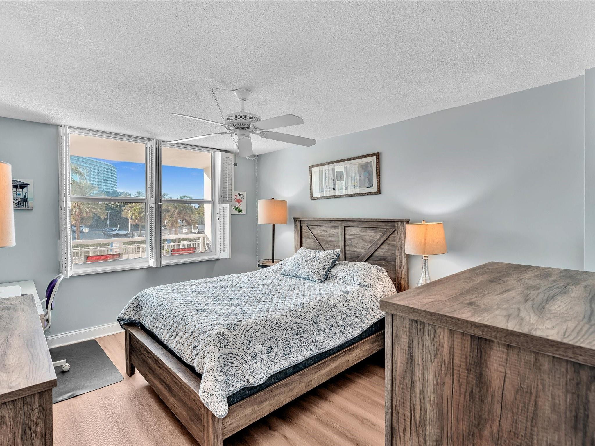 Bedroom with a wood bed and patterned white bedding beside windows overlooking palm trees and a high-rise.