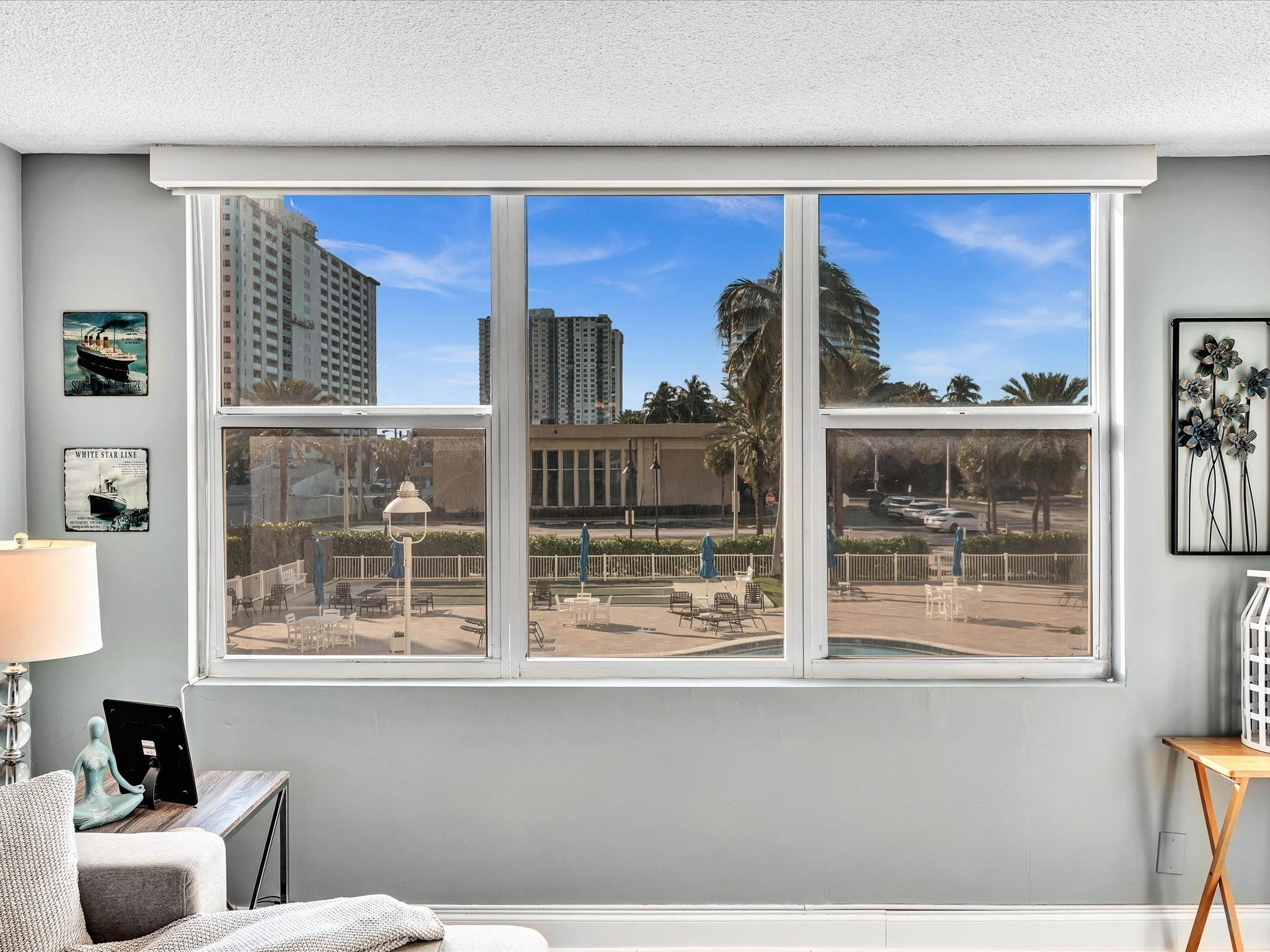 Living room with wide windows overlooking a pool deck, palm trees, and nearby high-rises.
