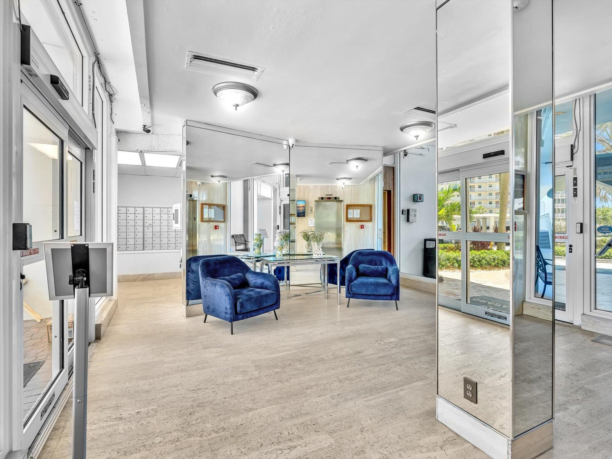 Condominium lobby with mirrored walls, mailboxes, and blue chairs beside glass entrance doors.
