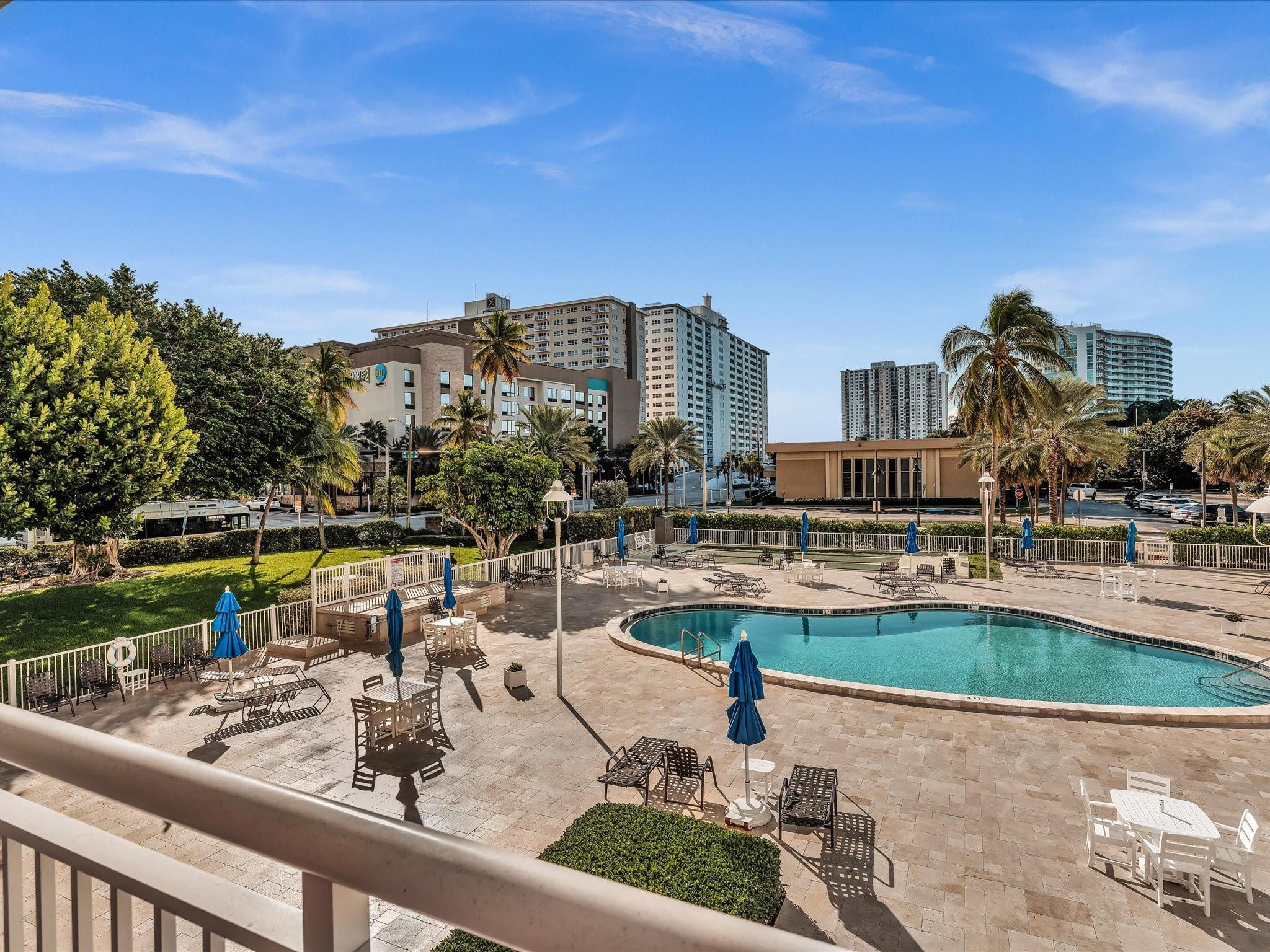 Outdoor pool deck with a curved pool, blue umbrellas, palm trees, and high-rise buildings.