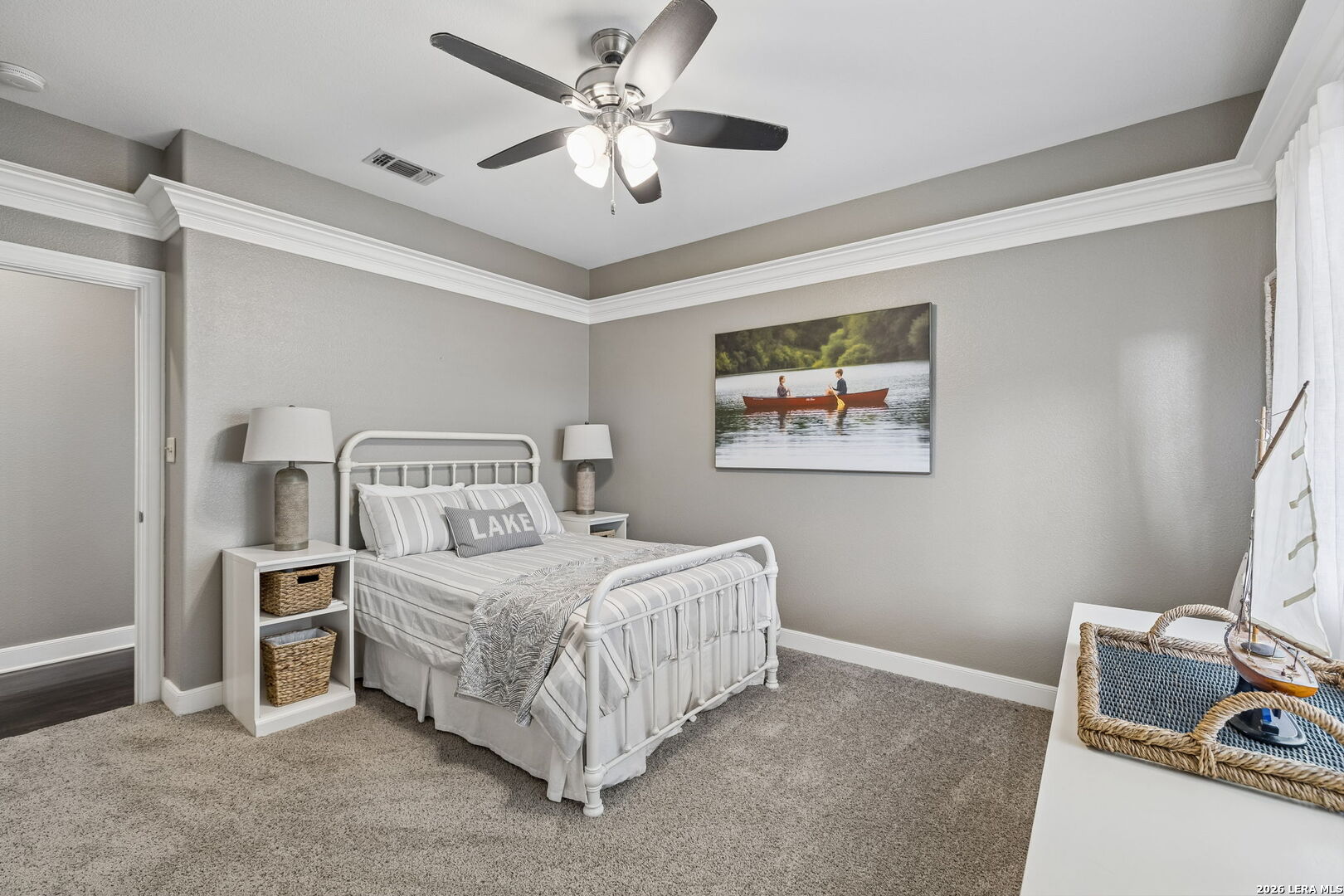 Staged bedroom with a white metal bed, striped bedding, lake-themed artwork, and a decorative sailboat.