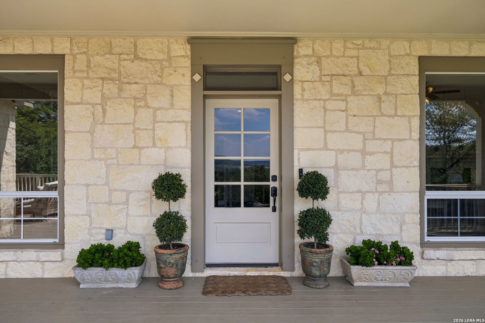 Stone home entrance with a white glass-paneled door, potted topiary, and flower planters.