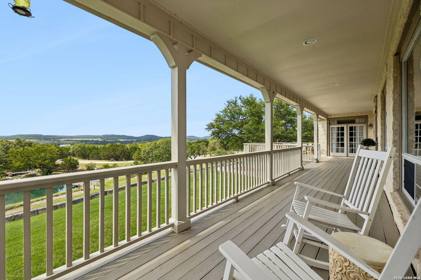 Covered porch with white rocking chairs overlooking green hills and a small patch of water.