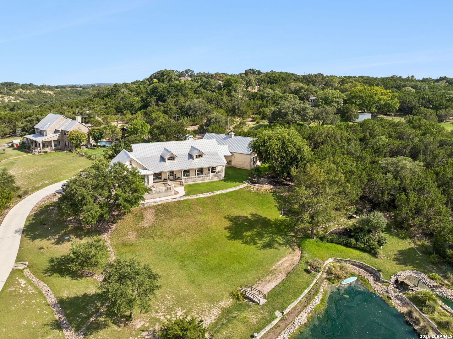 Aerial view of a stone home with a broad lawn, curved driveway, wooded acreage, and a small pond.